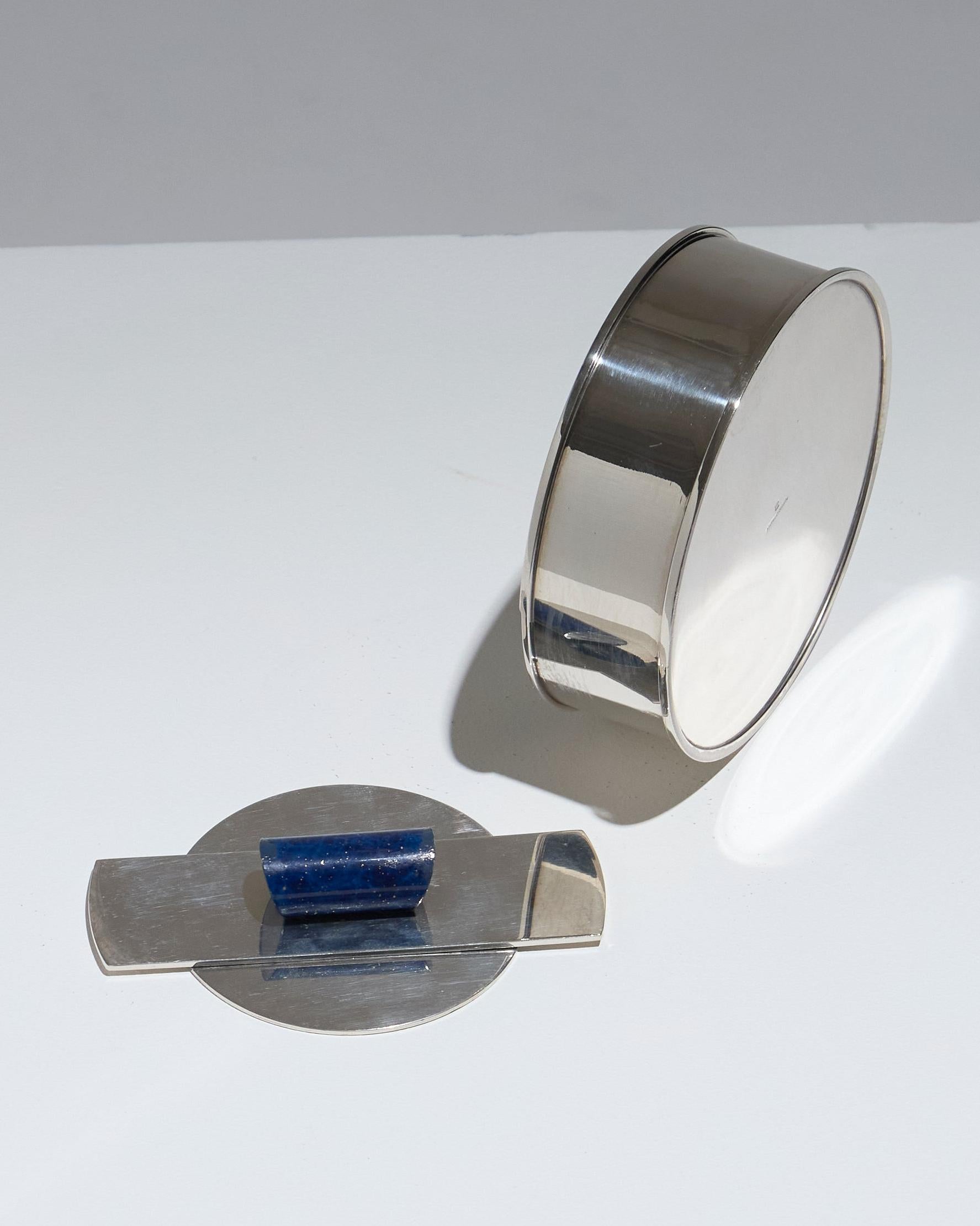 Boîte circulaire Art Déco en métal argenté, signée du poinçon de l'orfèvrerie française Puiforcat. Le couvercle géométrique est orné d'une poignée en résine bleu. En très bon état.