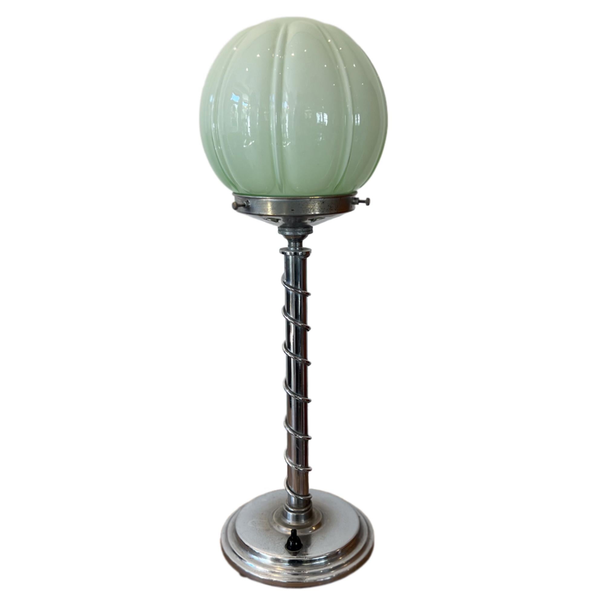 This decorative table lamp is made in a typical Art Deco design. 

With a chrome plated slim column, decorated with a twist of metal topped with an attractive opaque celadon green glass shade. When lit, the shade gives off a relaxing soft muted
