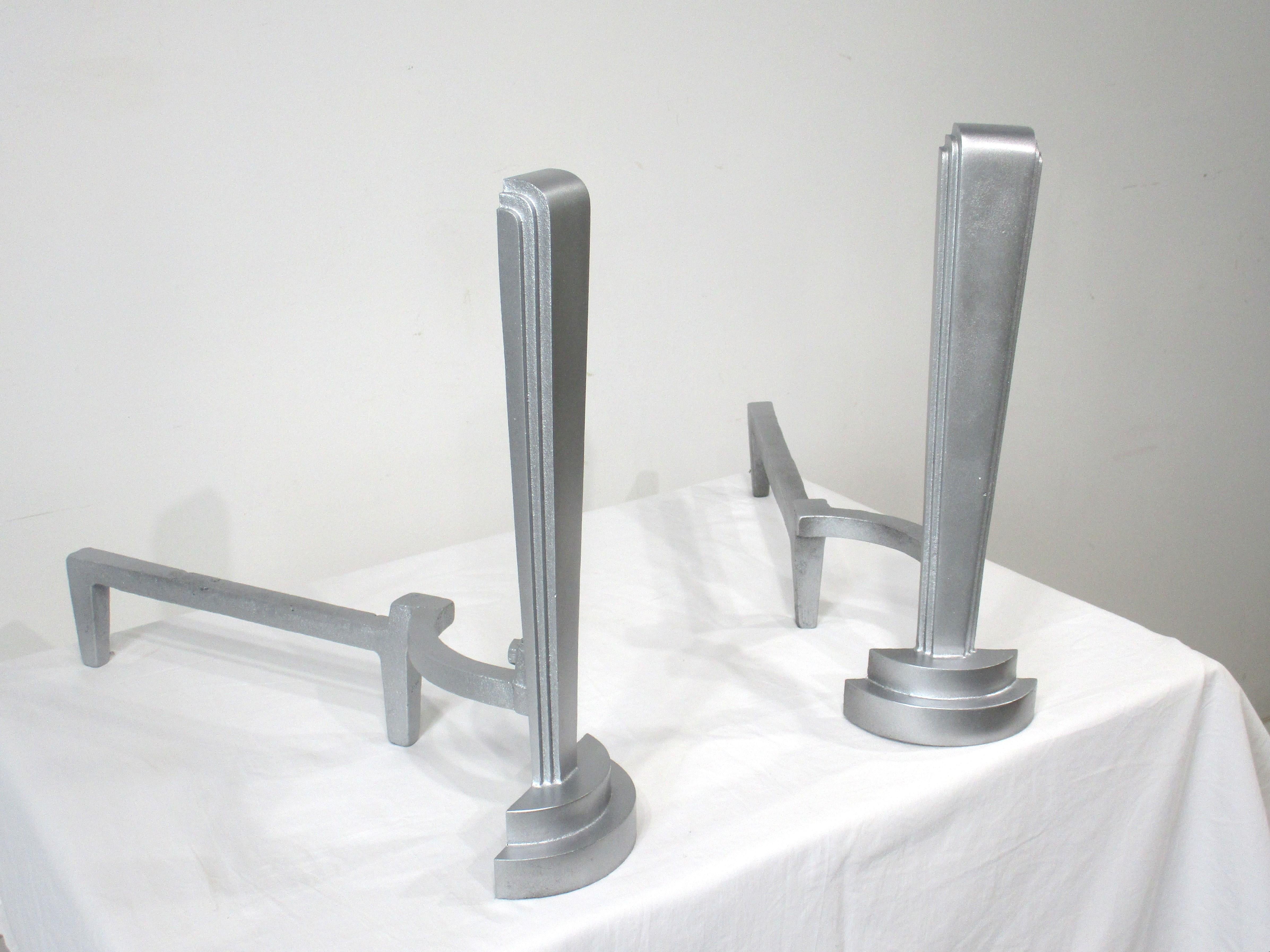 A pair of cast steel Art Deco styled fireplace andirons having a painted silver gray high temperature coating . A very simple design with graduating lines and form in the manner of the skyscraper style in the early 1930's and 40's . Marked to the