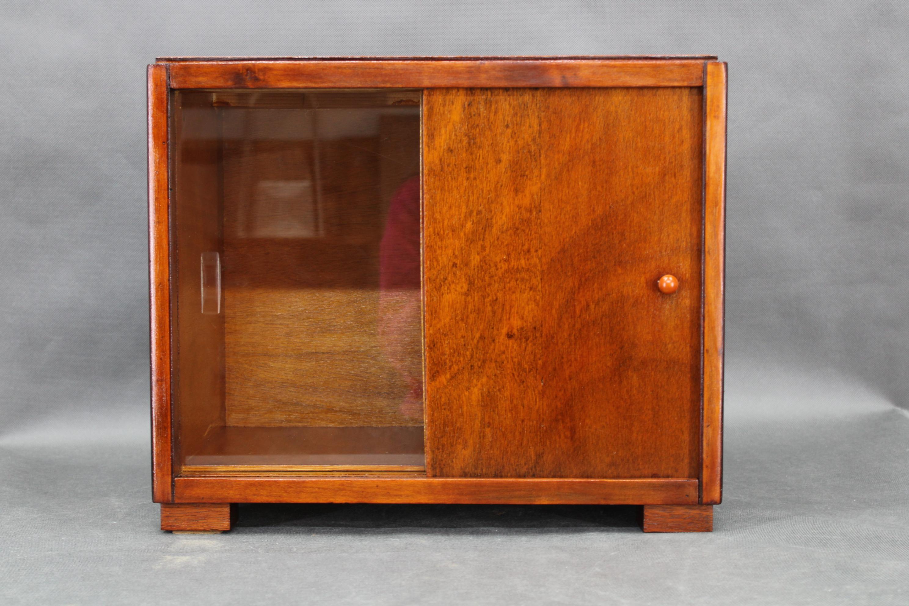 Diese kleine Art-Déco-Vitrine wurde in den 1940er Jahren in der Tschechoslowakei hergestellt. Das aus Buchensperrholz und Fichtenkernplatte gefertigte Möbelstück mit Glastür weist klare geometrische Linien auf, die für die Formensprache der späten