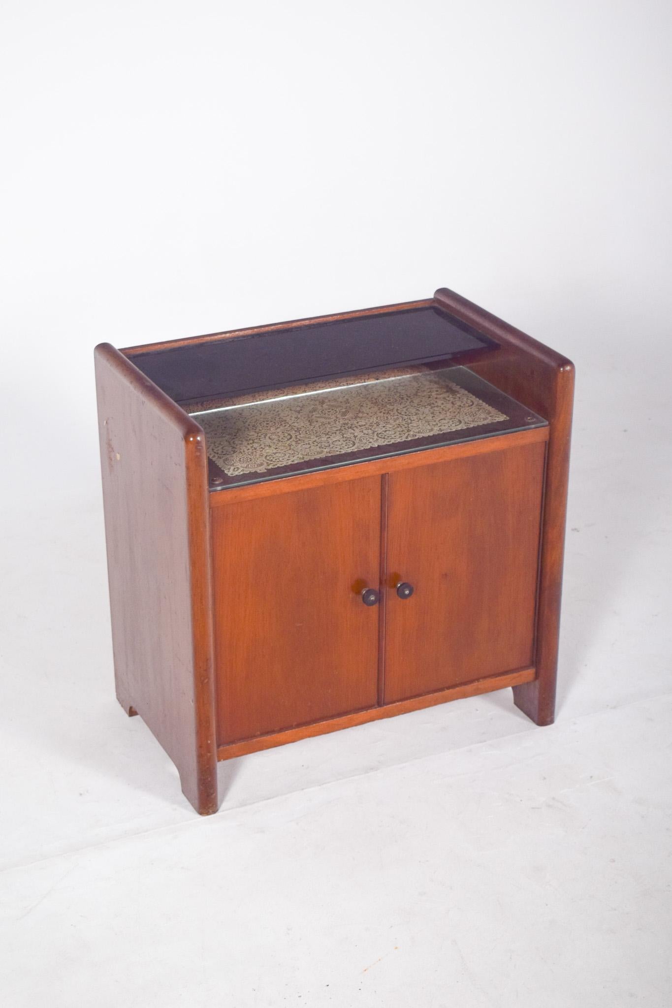 Petit meuble de bar à deux portes. La partie supérieure est constituée d'un plateau en verre foncé et, juste en dessous, d'une zone d'exposition également en verre, dont le fond est tapissé de papier décoratif. Cette zone est idéale pour placer des