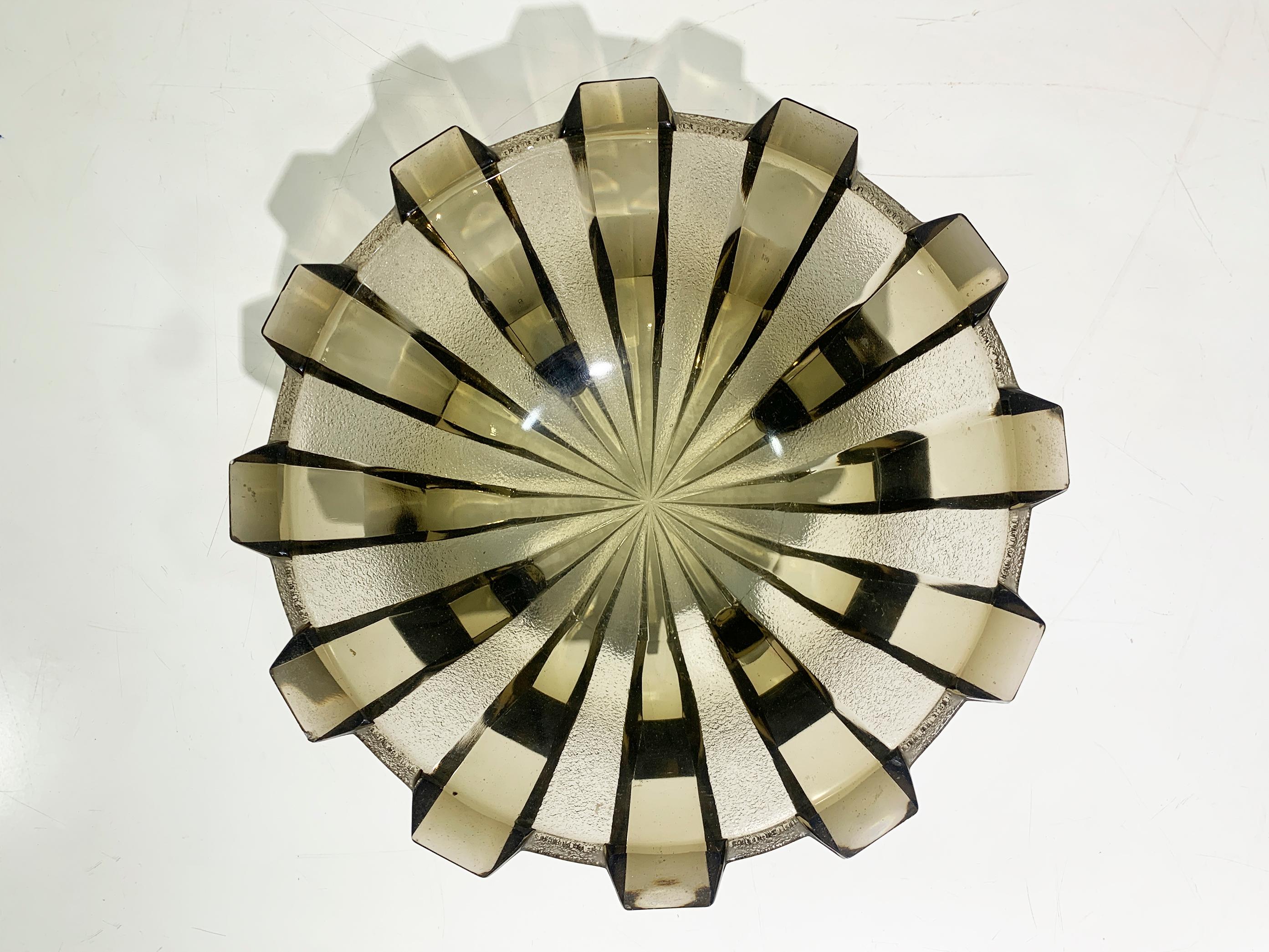 Este cuenco, de la década de 1930, muestra el atrevido estilo geométrico y la artesanía característicos del diseño Art Déco. El patrón estriado y facetado que irradia desde el centro le da un aspecto llamativo, casi escultórico, y refleja la luz