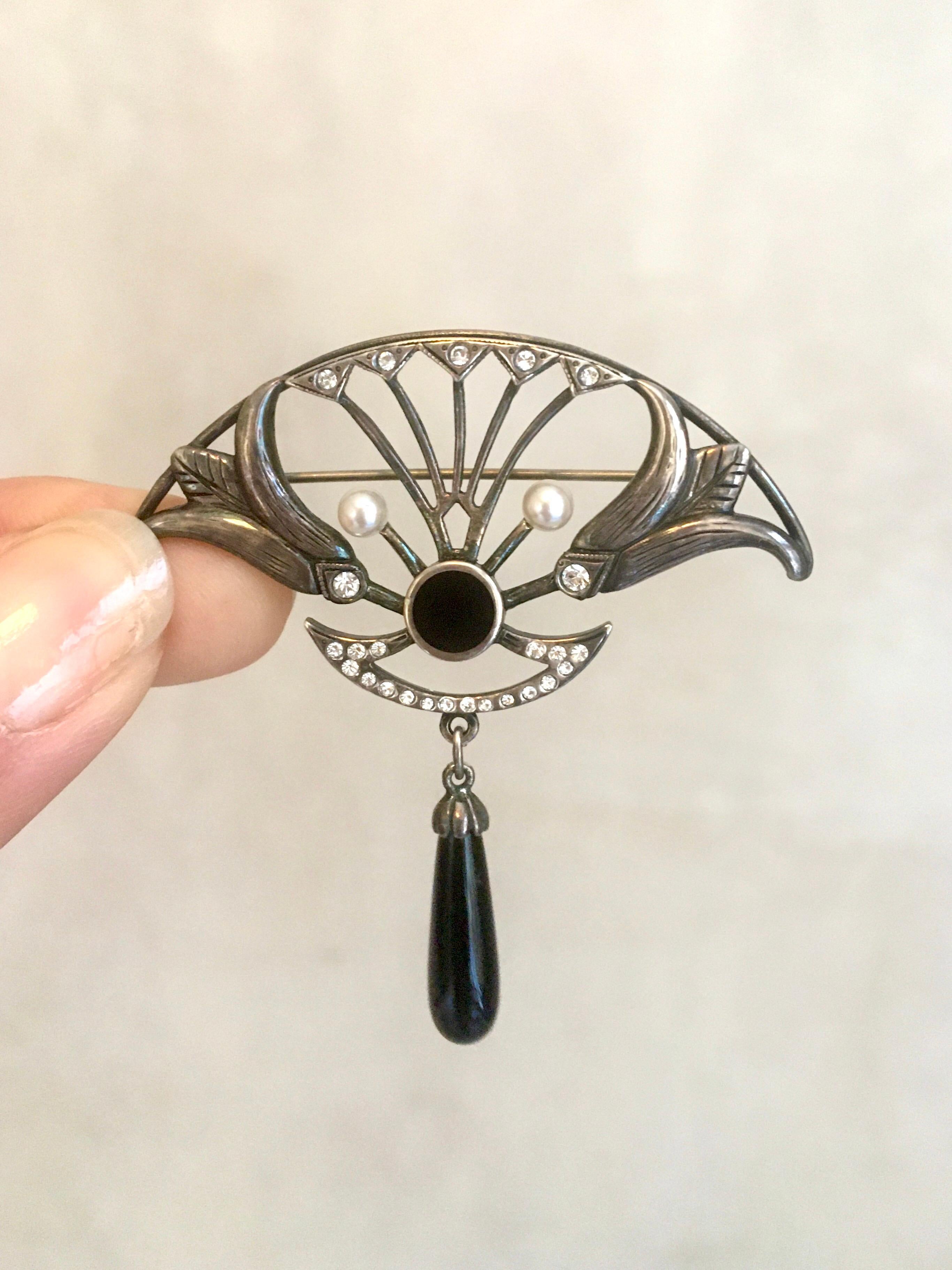 An Art Deco sterling silver brooch set with black onyx and pearls. The silver openwork top is set with shimmering rows of crystal diamonds and two white cultured pearls. The silver has carved flower accents, which provides a wonderful texture to the