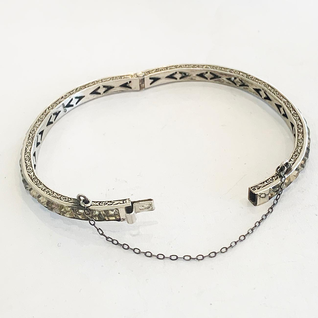 Art Deco Bangle in Sterling Silver carved to both sides in a fine Flower and Leaf decoration, Channel set with white Paste Gems to the whole perimeter, with original clasp and Safety Chain. All in excellent order with no damage or losses, soft age