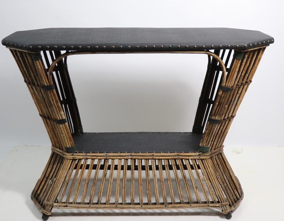 Unusual form stick wicker console table with textured black vinyl covered top and lower shelf. The top is eight sided, the vinyl top is held in place with brass studs, the lower shelf ( 10 in H ) is also black vinyl. The wicker shows some nicks and
