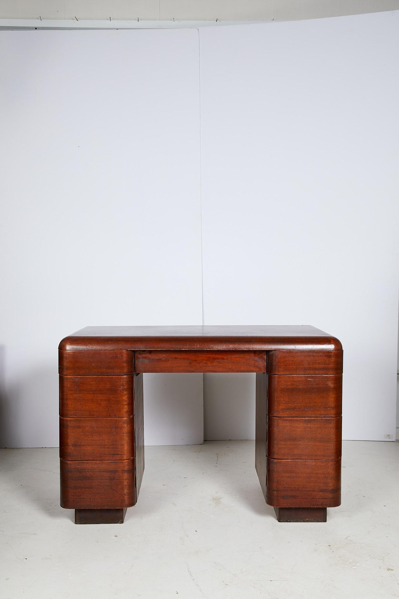 The 20th century streamlined writing desk was designed by Paul Goldman in 1946 for Plymold Co. in the Art Deco style. The bentwood desk holds a shallow center drawer flanked by pedestals with eight graduated drawers that are flushed into the case