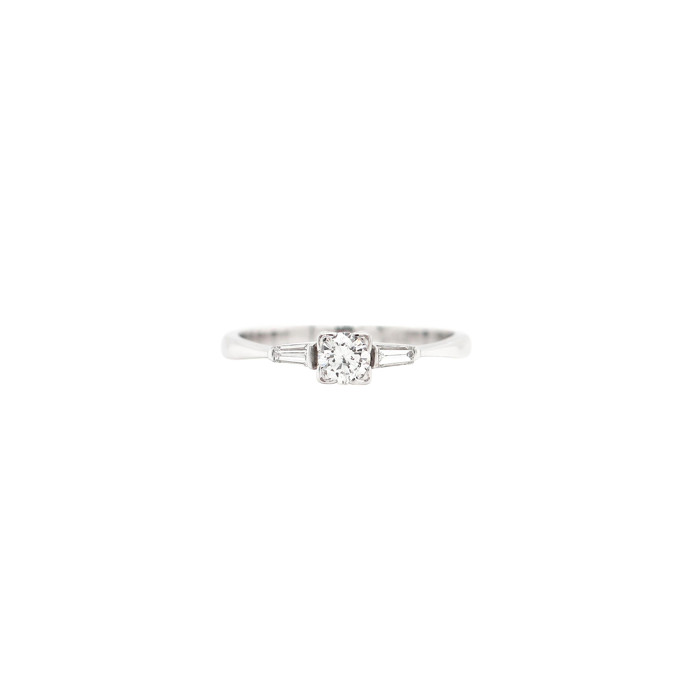 Ein schöner Verlobungsring im Art-Deco-Stil, besetzt mit einem runden Diamanten im Brillantschliff mit einem Gewicht von 0,30 ct in einer offenen Kastenfassung. Der Diamant in der Mitte wird von zwei spitz zulaufenden Diamanten im Baguetteschliff