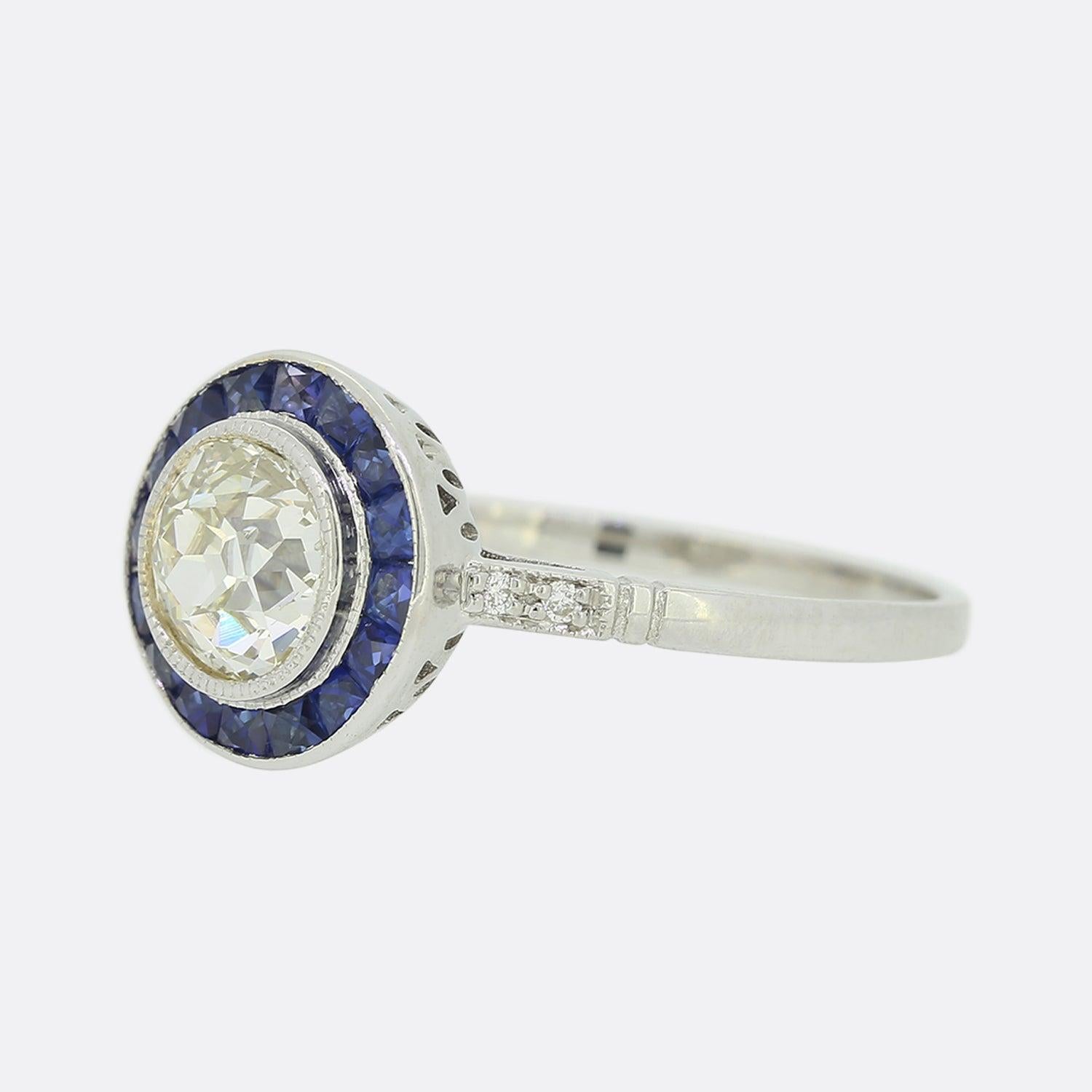 This is an Art Deco style 18ct white gold, sapphire and diamond ring. The focal point of the ring is a 1.0 carat old cut diamond, in a rub-over milgrain setting. Surrounding the centre stone are multiple calibrated sapphires, which are well matched