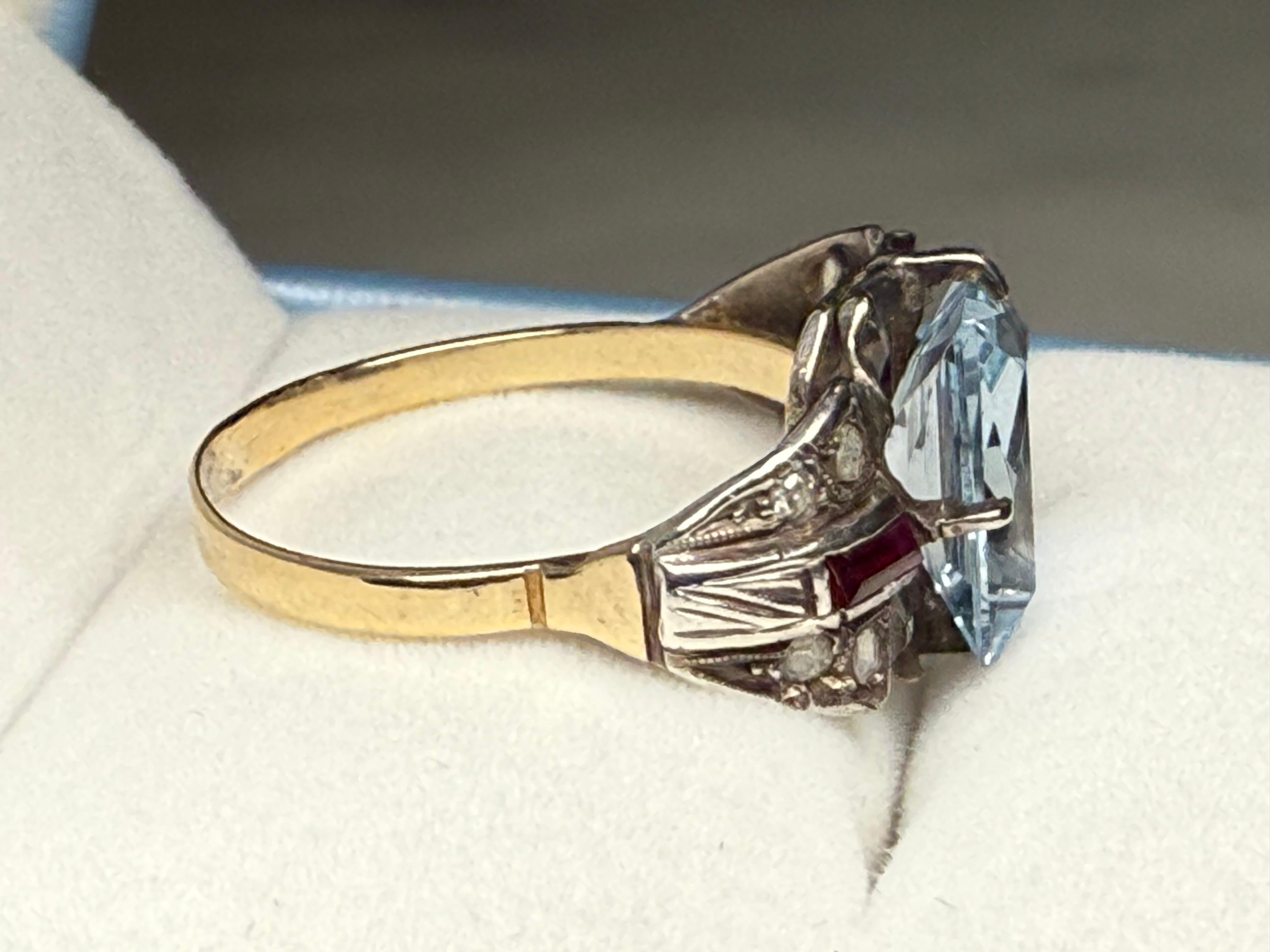 Si tratta di un anello in stile Art Deco a 14 carati. Raffigura una pietra di acquamarina di forma rettangolare con incastonatura a forchetta, ornata su ciascun lato da un ventaglio in oro bianco arricchito da rubini e diamanti con incastonatura a