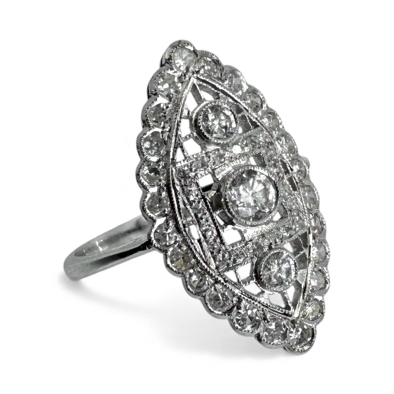 Cette bague à diamants de style Art déco en or blanc 14 carats présente des détails ajourés et milgrainés sur la face allongée en forme de navette. Le design est centré sur trois diamants ronds de taille brillant sertis en lunette, totalisant