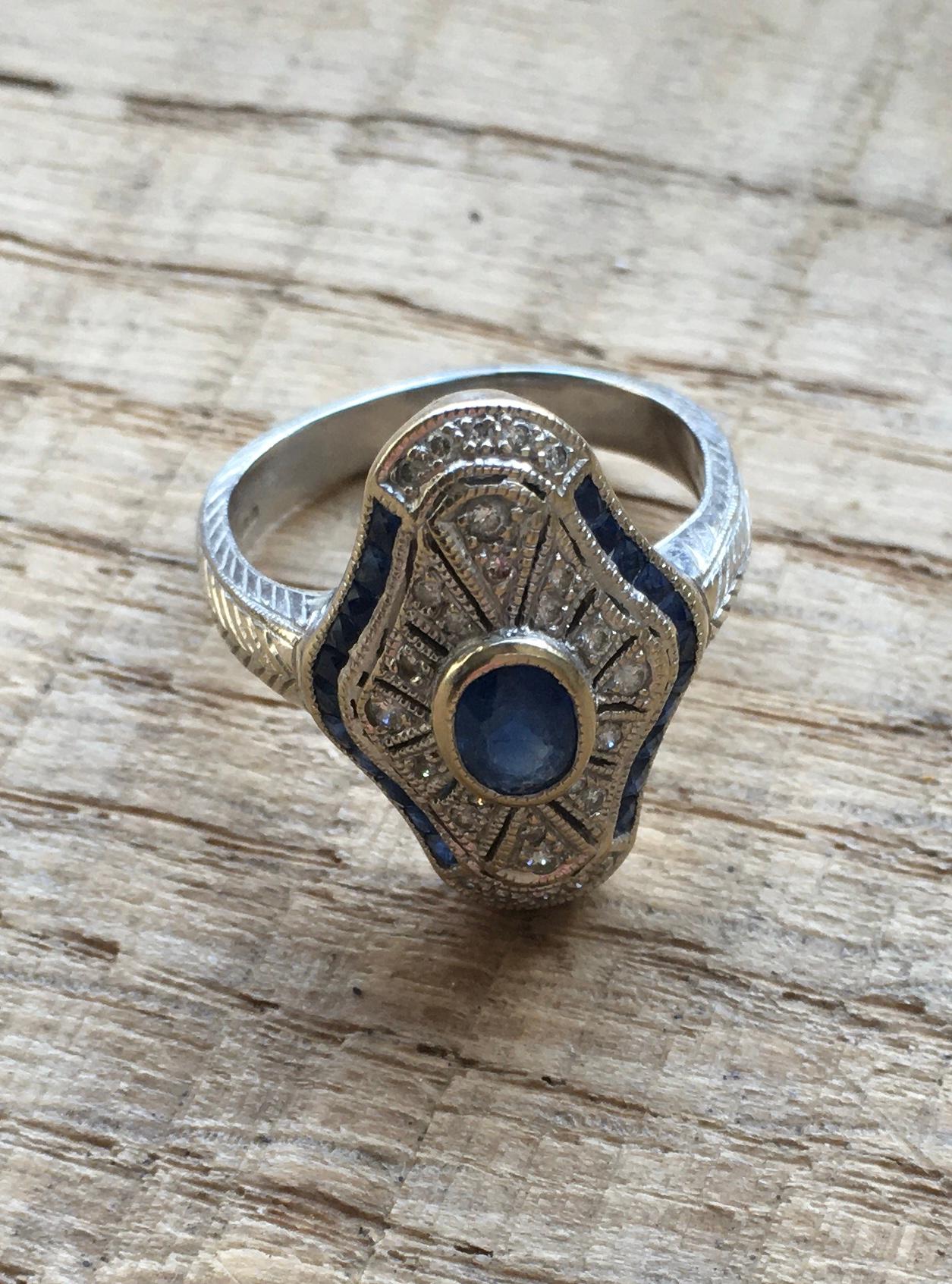 A hallmarked 18ct gold white gold, sapphire and diamond Art Deco style panel ring. The ring being bezel set with an oval cut sapphire with round cut diamond accent stones and two rows of sapphire accent stones to the head. The shoulders having