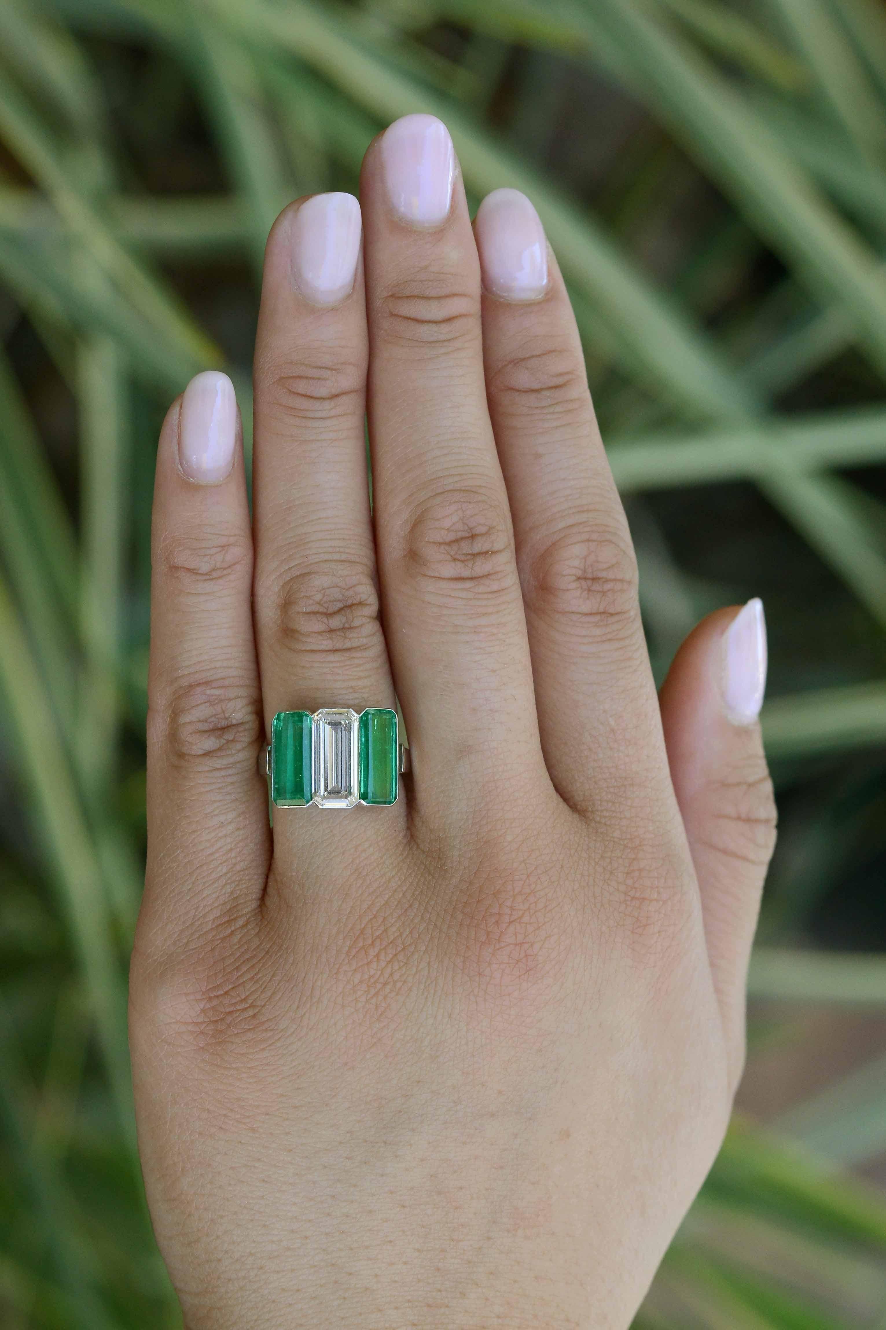A premiere three stone diamond and emerald engagement ring, hand fabricated in a sleek Art Deco style. At it's center, an exquisite 3 carat emerald cut diamond is shouldered by two vivid green Colombian emeralds, creating prosperous symmetry and