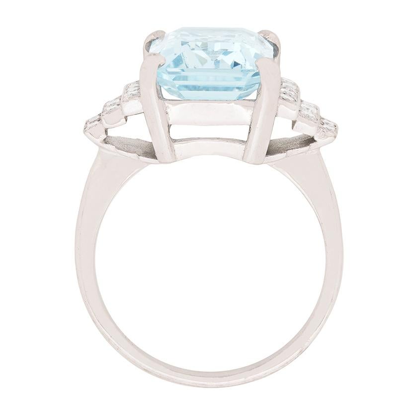 This glamorous dress ring, which gets its inspiration from the Roaring Twenties, presents a placid blue, 5.60 carat, emerald cut aquamarine between a pair of stepped, diamond set shoulders in 18 carat white gold. The ring's geometric shoulders,