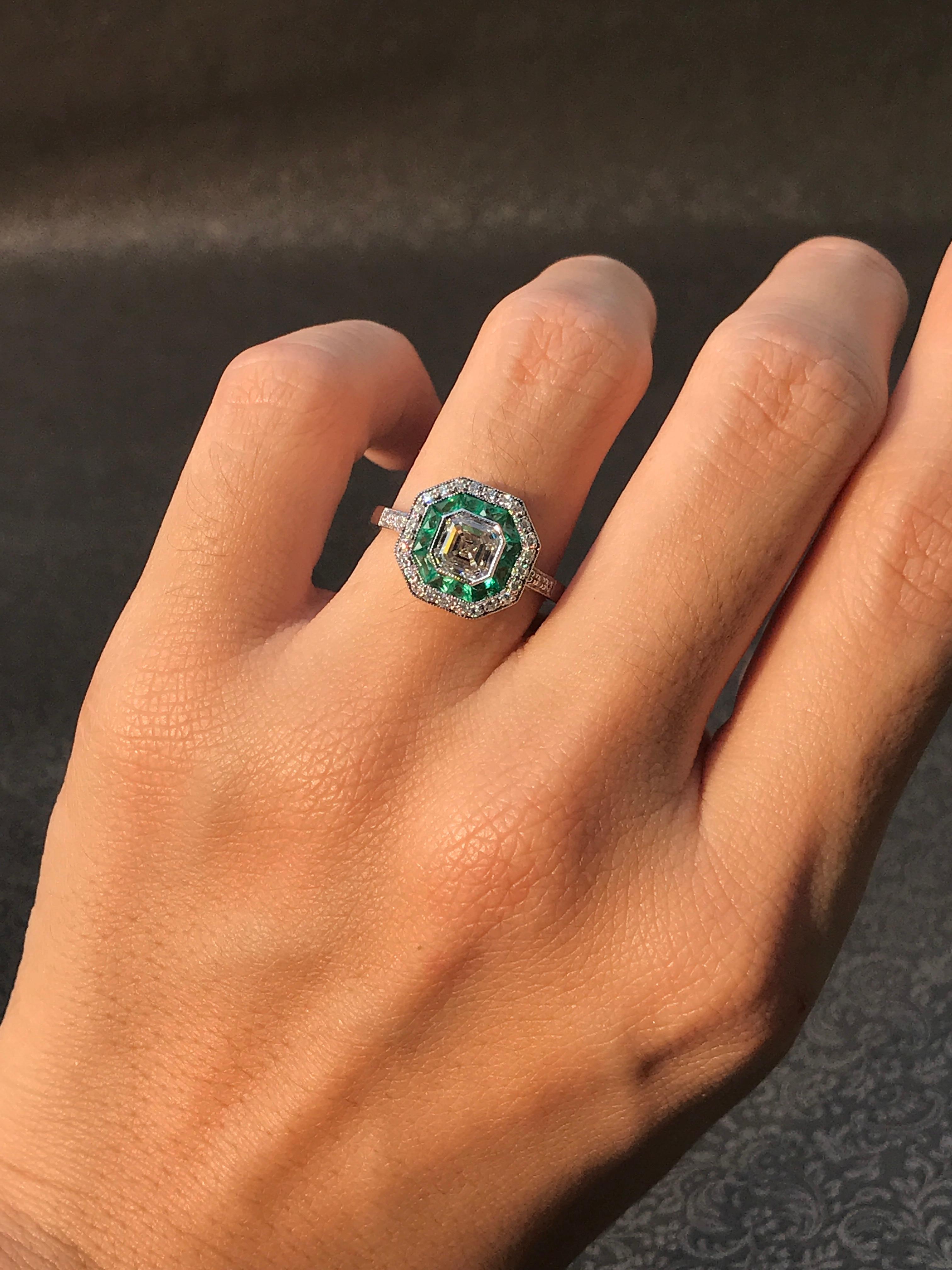 An 18k white gold Art-Deco design diamond and sapphire target ring, center asscher cut K/VS diamond with an inner halo of French cut emeralds and outer round diamonds with finished with millgrain edging, finished with diamond shoulders.

Ring