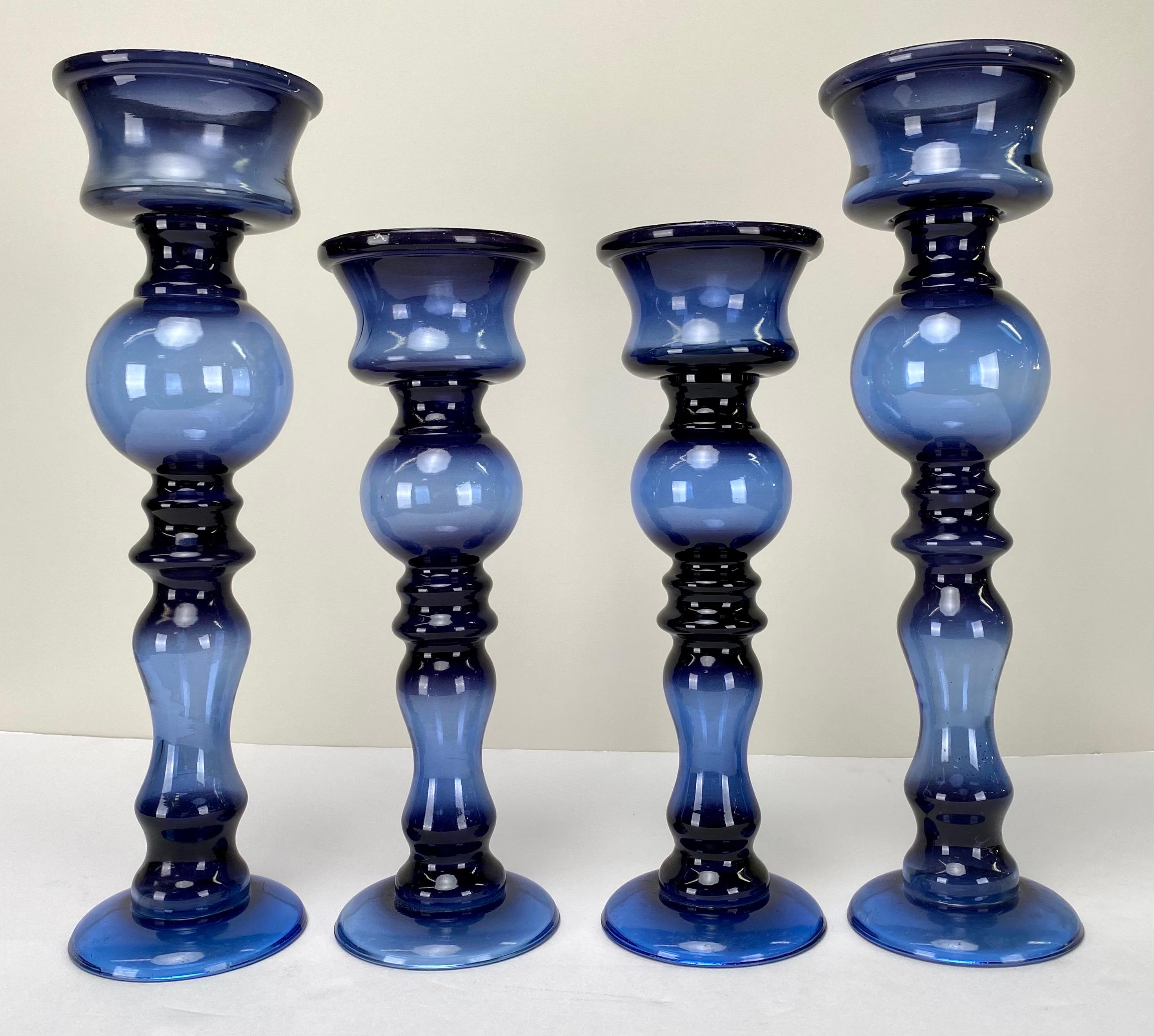 A set of four Art Deco-inspired Blue Candle Holders, bathed in a rich, dark blue hue. This enchanting set comprises two grandiose candle holders alongside two smaller counterparts. Each piece boasts a stem adorned with artfully arranged bubbles,