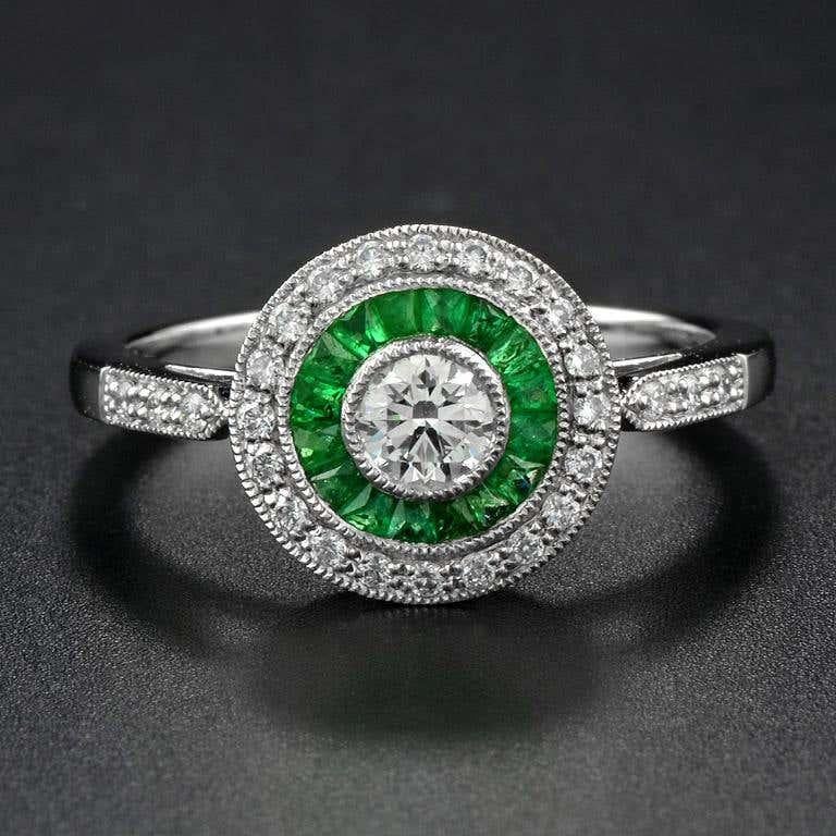 This Art-Deco engagement ring is completely spectacular! The vibrant green emeralds are a specialty cut to surround the excellent round brilliant cut center diamond, which is in a thin bezel with mil-grain detail. Crafted in Platinum.
Ring
