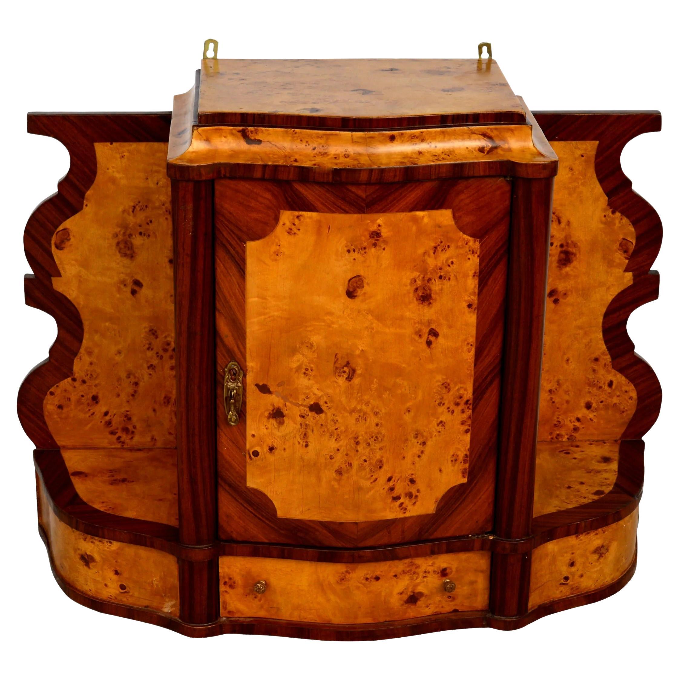 Hanging wall apothecary cabinet in a blend of exotic burl wood featuring scrolled sides with rosewood inset borders. One door opens to two shelves with a drawer underneath with a nickel pull.
 The cabinet hangs from hardware secured to the back of