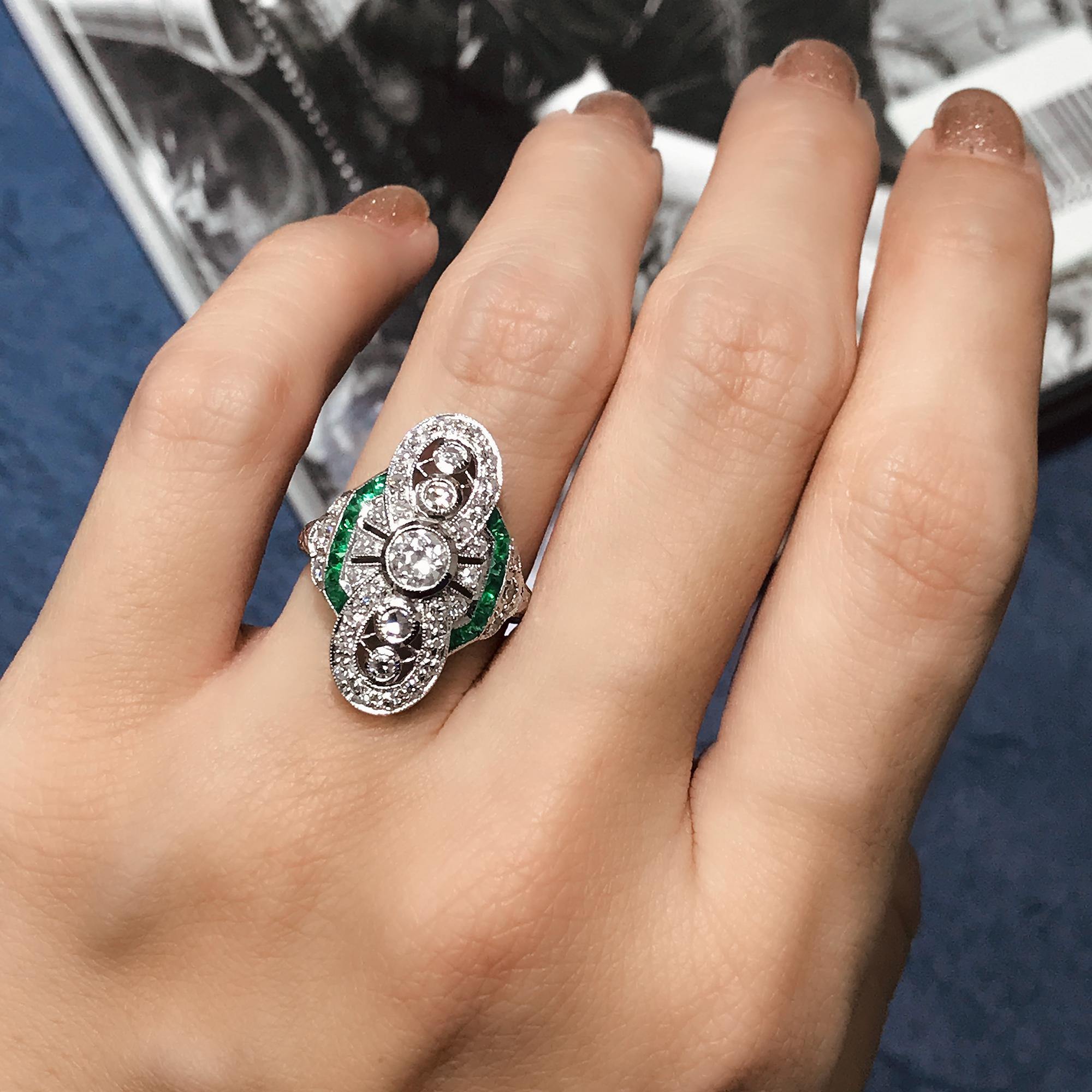 This Art Deco style ring sits low and hugs the finger nicely. It features round old cut diamonds in the center with French cut emerald on its west and east. Then multiple round cut diamond surround it. The milgrain adds to the vintage style.  

Ring