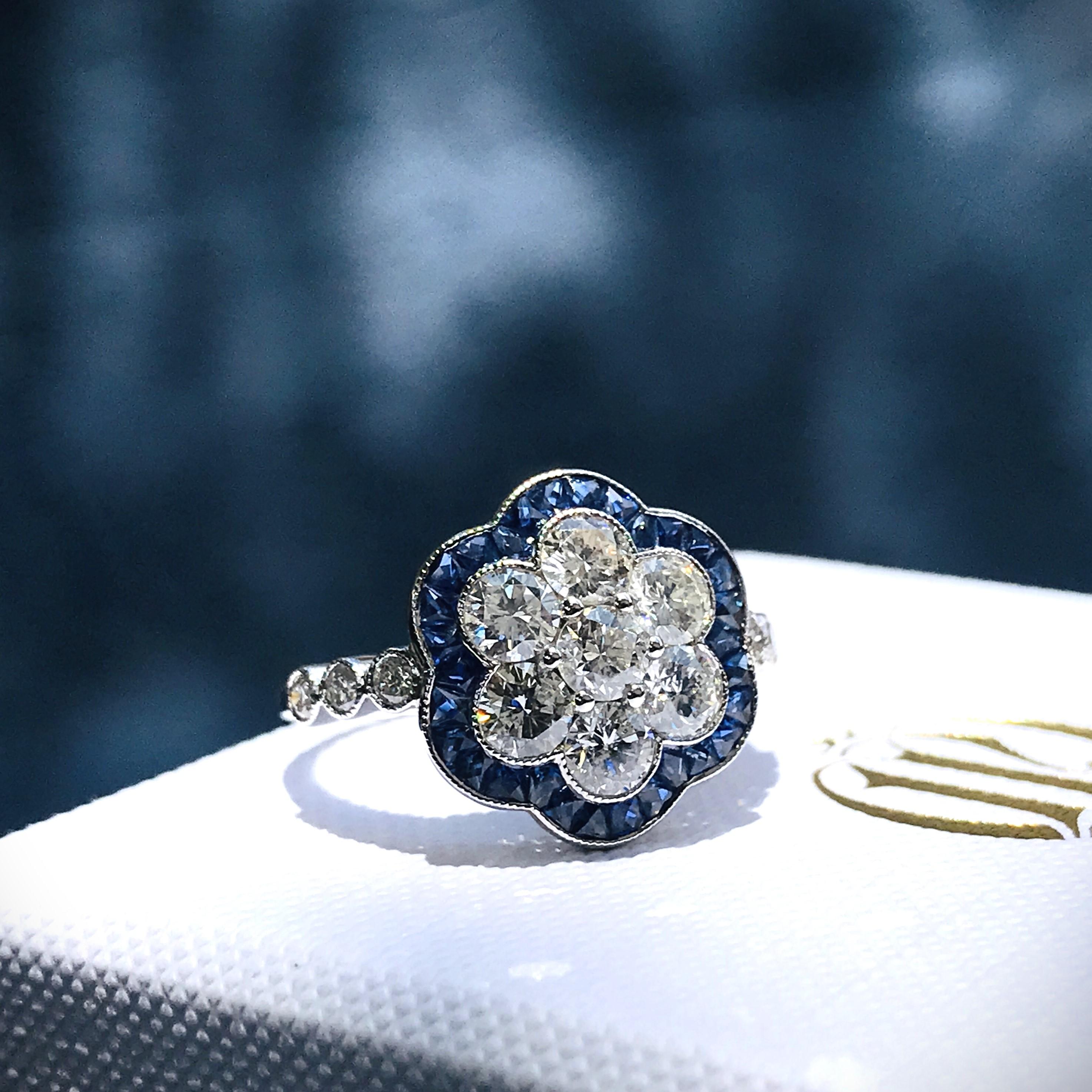 Artistry and sophistication come together in a flower ring with sapphires and diamonds from the Art Deco inspiration. The ring features a round shape center diamond on it surrounded by French Cut Sapphires, round brilliant diamonds on shoulders add
