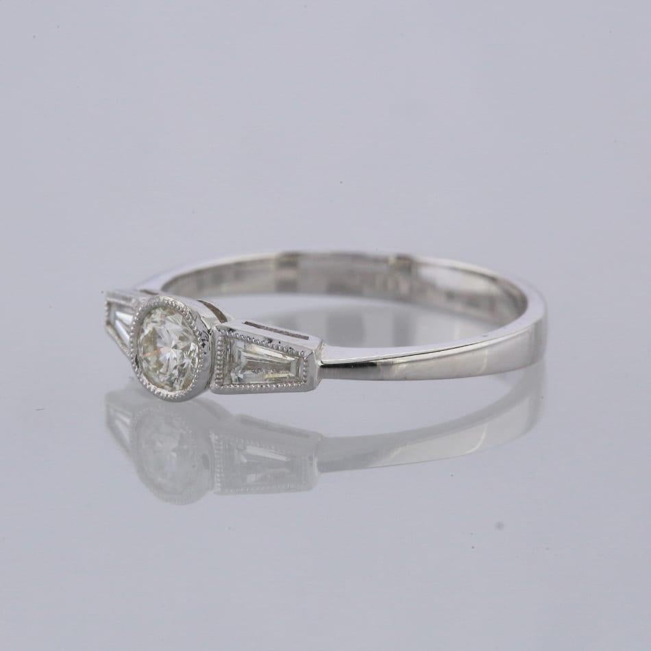 This is an 18ct white gold diamond ring. The central round brilliant cut diamond is flanked by 2 beautiful tapered baguette cut diamonds in an art deco style.

Condition: Used (Very Good)
Weight: 2.5 grams
Size: O
Total Diamond Carat Weight: Approx.