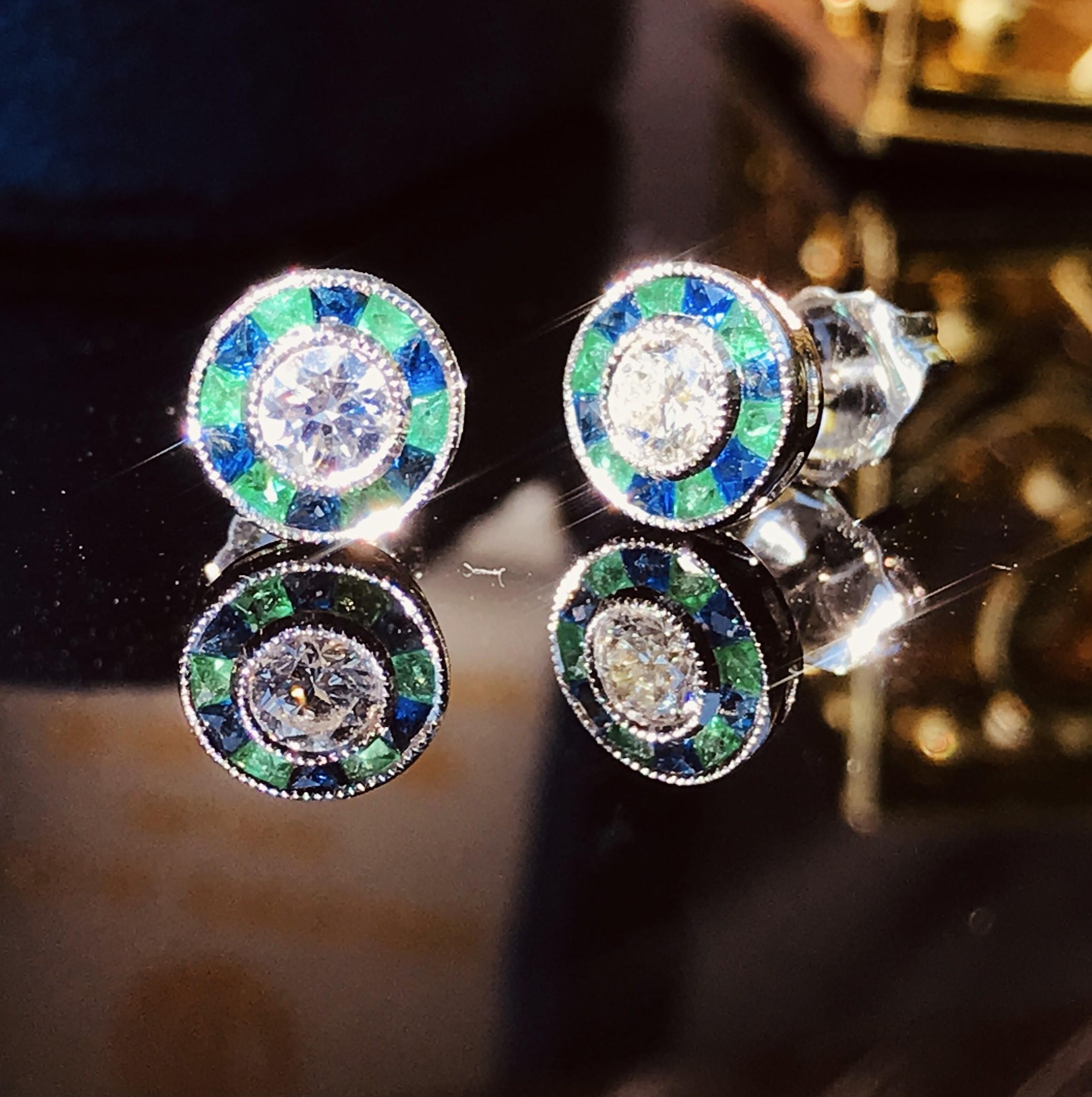 A diamond with emerald and sapphire target earrings, rub-over-set to the center with a round brilliant cut diamond surrounded by French cut emeralds and sapphires, all set in 18k white gold. A gorgeous diamond with emerald and sapphire target
