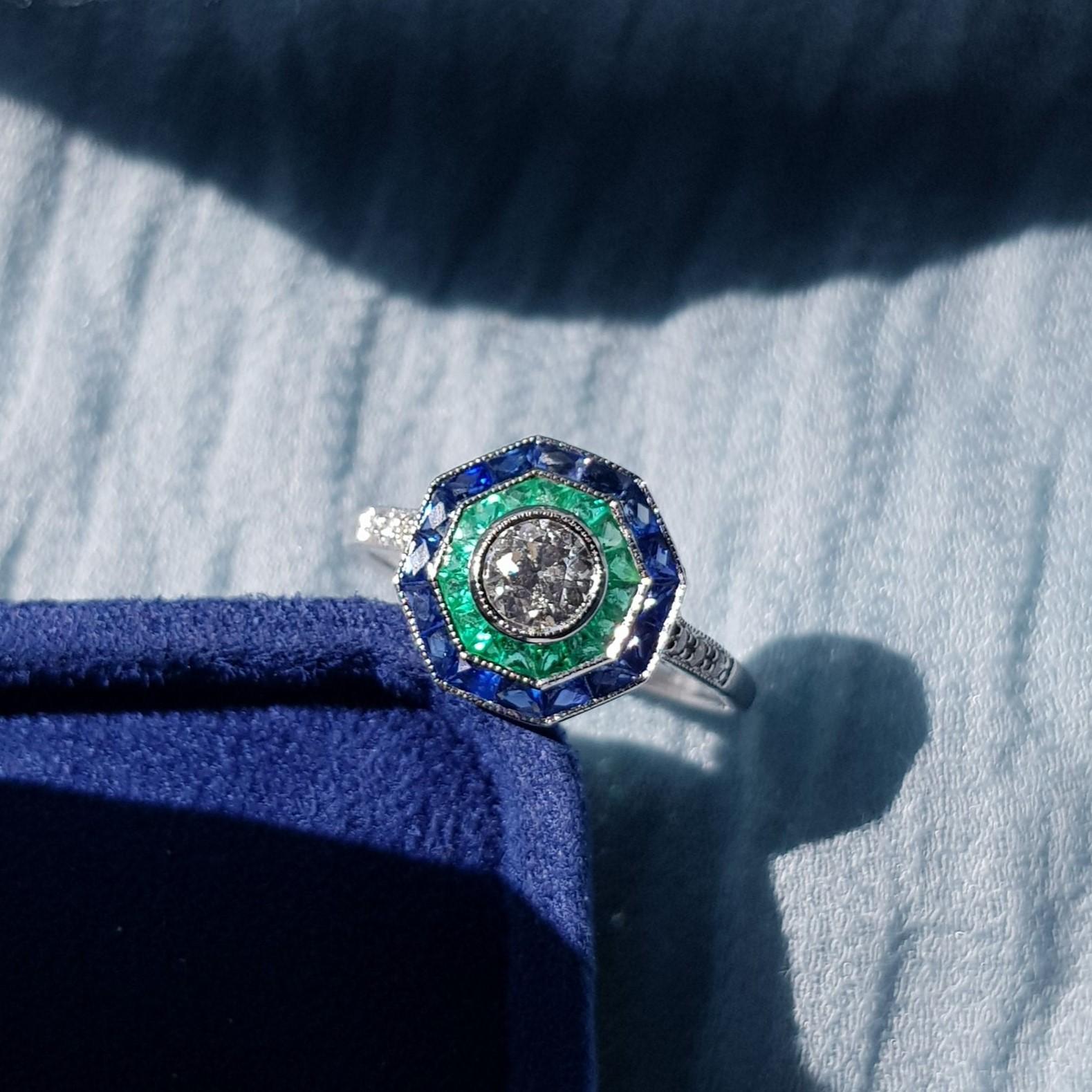 An incredibly stylish Art Deco target ring, set with a double halo clusters of gemstones – blue sapphire  and emerald around a central old cut diamond. The diamonds are bright, displaying excellent flash and sparkle, while the sapphire and emerald