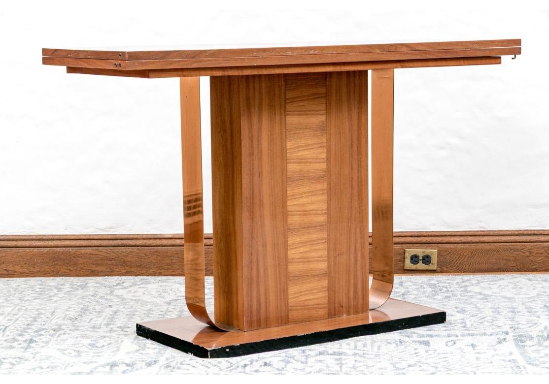 An Art Deco style games table in warm medium brown wood, showcasing a long rectangular table top that pulls open, flips to a spacious table surface and latches when not in use. The tabletop is raised over an impactful pedestal base with a wide