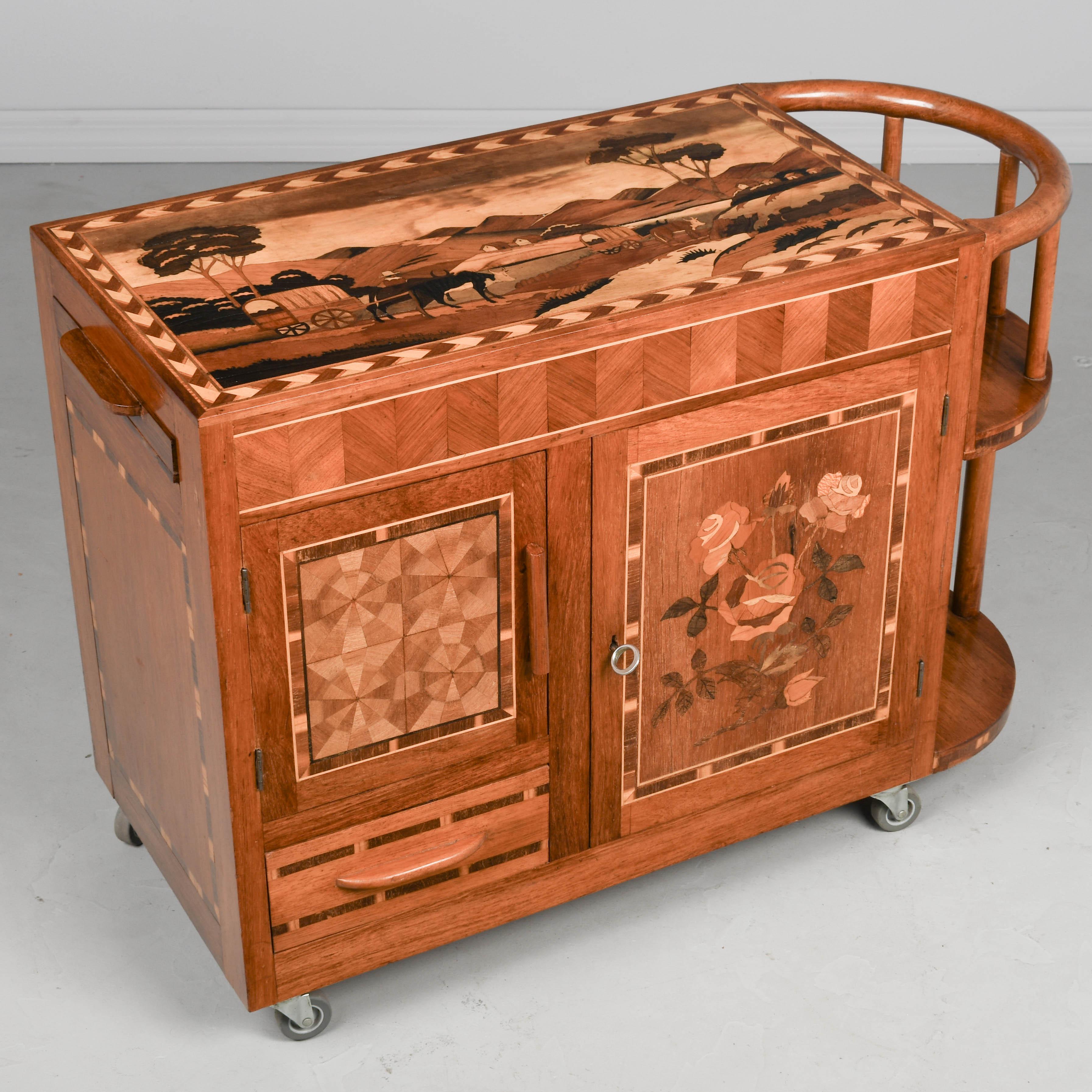 Ein italienischer Bar- oder Getränkewagen im Art-Déco-Stil mit Intarsien aus Mahagoni, der rundherum reichhaltig mit verschiedenen Holzeinlagen verziert ist. Der obere Teil zeigt eine pastorale Szene mit von Rindern gezogenen Planwagen.
