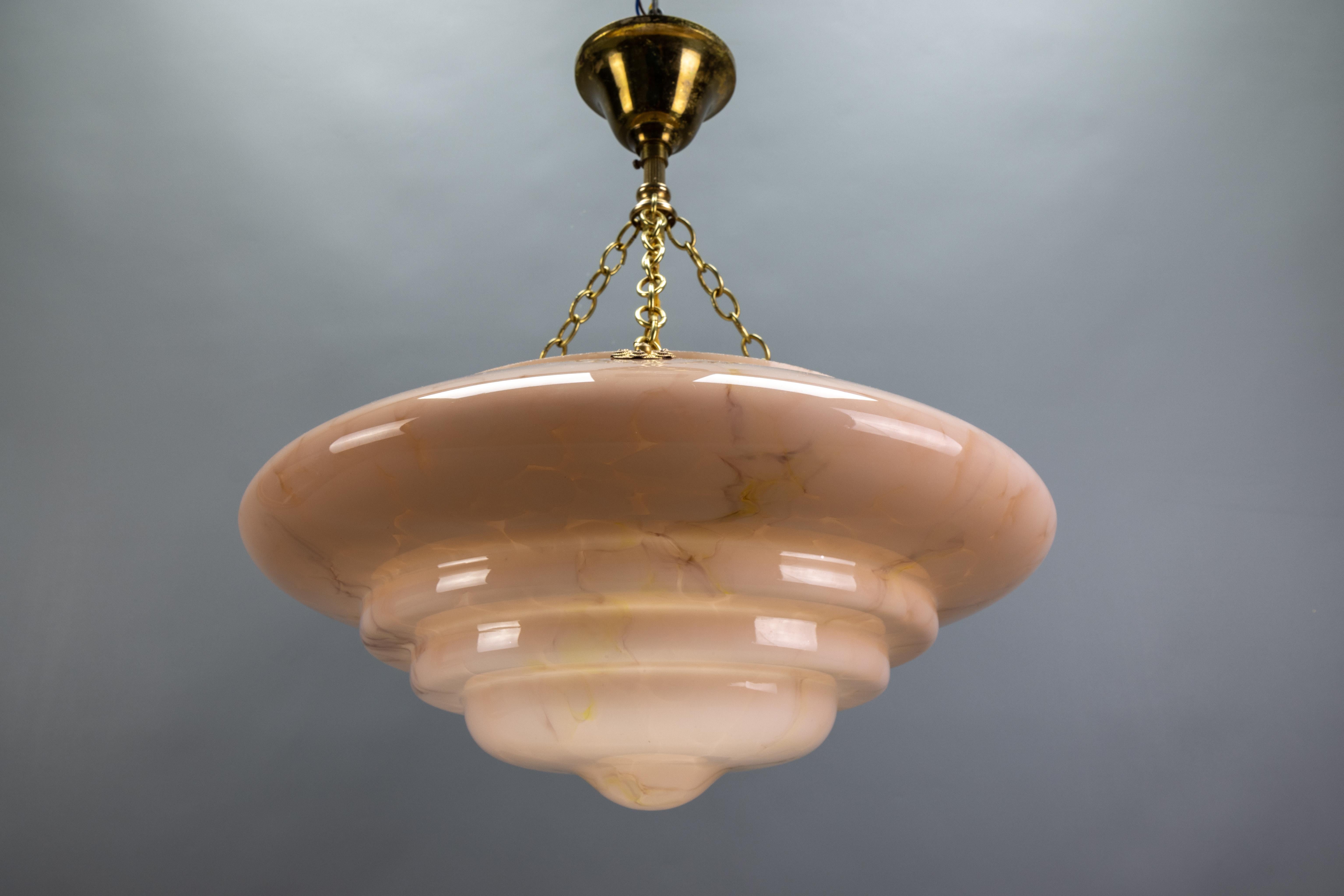 An Art Deco style marbled layered beige pastel pink glass and brass pendant light from circa the 1950s, Germany.
This beautiful ceiling light fixture features a marbled and layered glass lampshade in beige pastel pink and light brown tones. The
