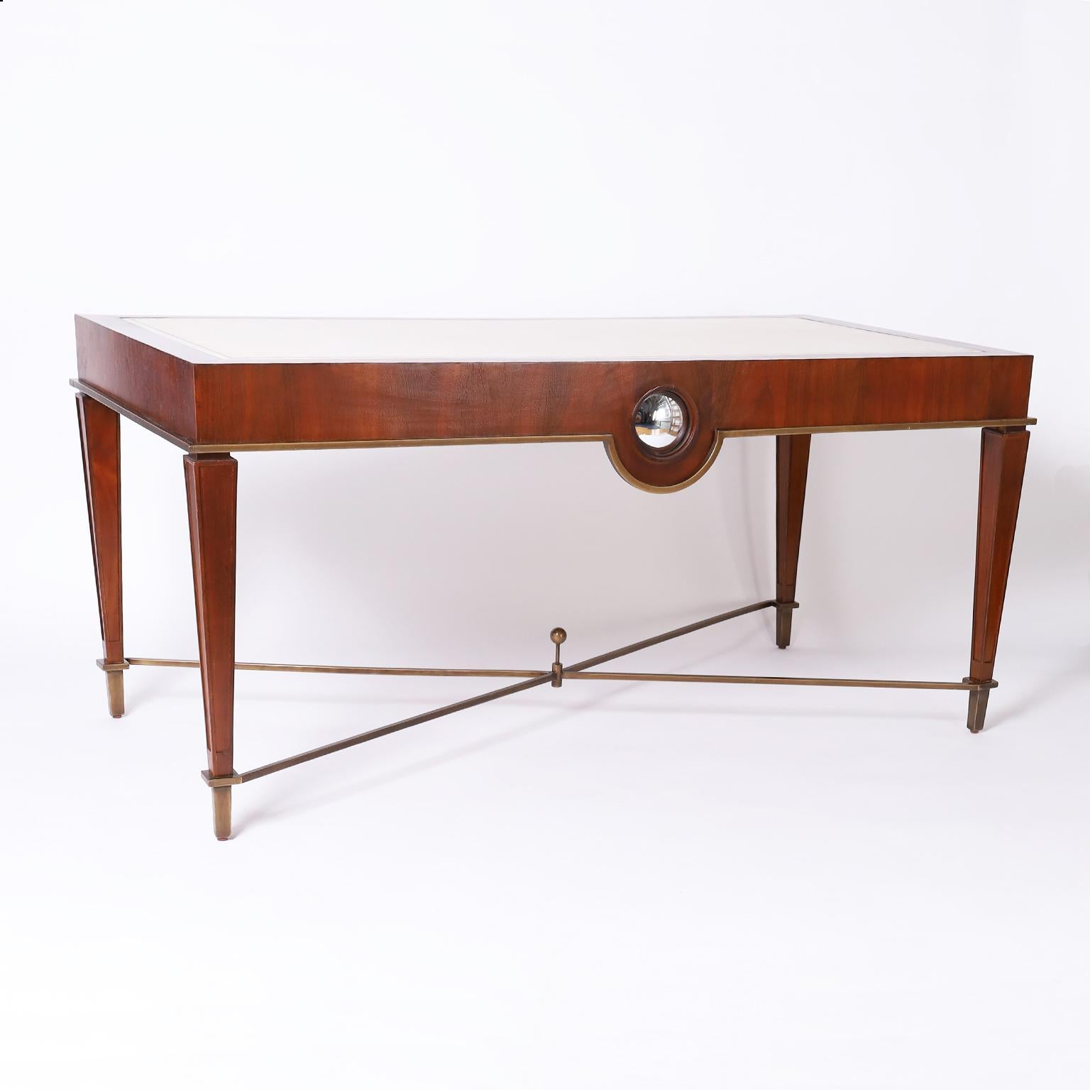 Three drawer desk or writing table crafted in mahogany with a white leather top over a sleek elegant form with art deco influence, finished all around with a convex mirror on the back and a bronze stretcher with center ball and attached feet.