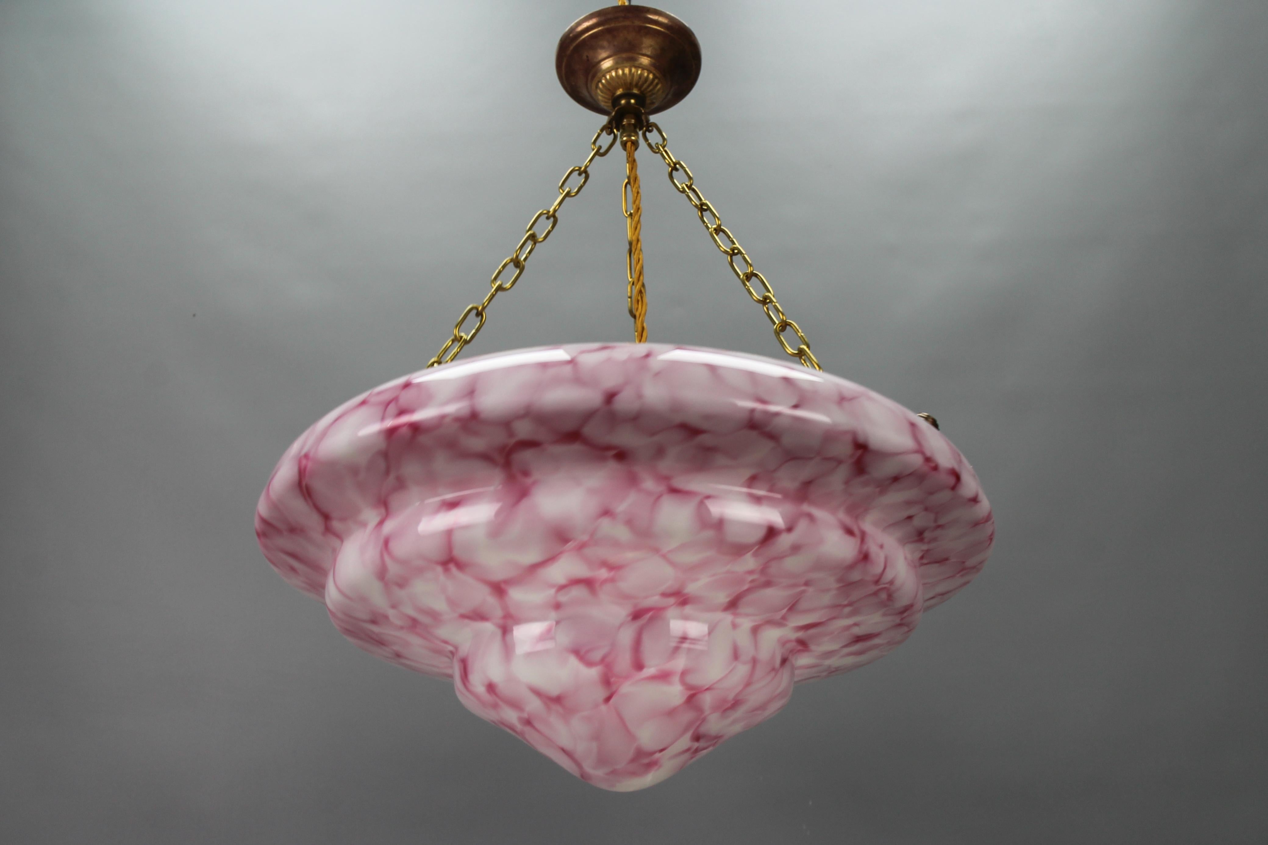 An Art Deco style marbled layered pink and white color glass and brass pendant light from circa the 1950s, Germany.
This beautiful ceiling light fixture features a marbled and layered glass lampshade in pink and white. The glass bowl is hung on