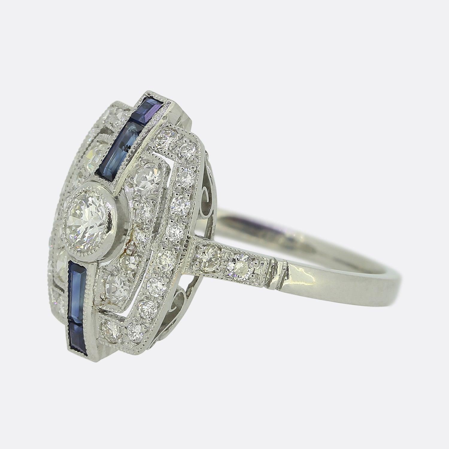 Ecco un favoloso anello a grappolo con zaffiri e diamanti che rende omaggio in modo eccellente a uno stile Art Deco senza tempo. Un singolo diamante rotondo taglio brillante si trova leggermente rialzato al centro del quadrante in una raffinata