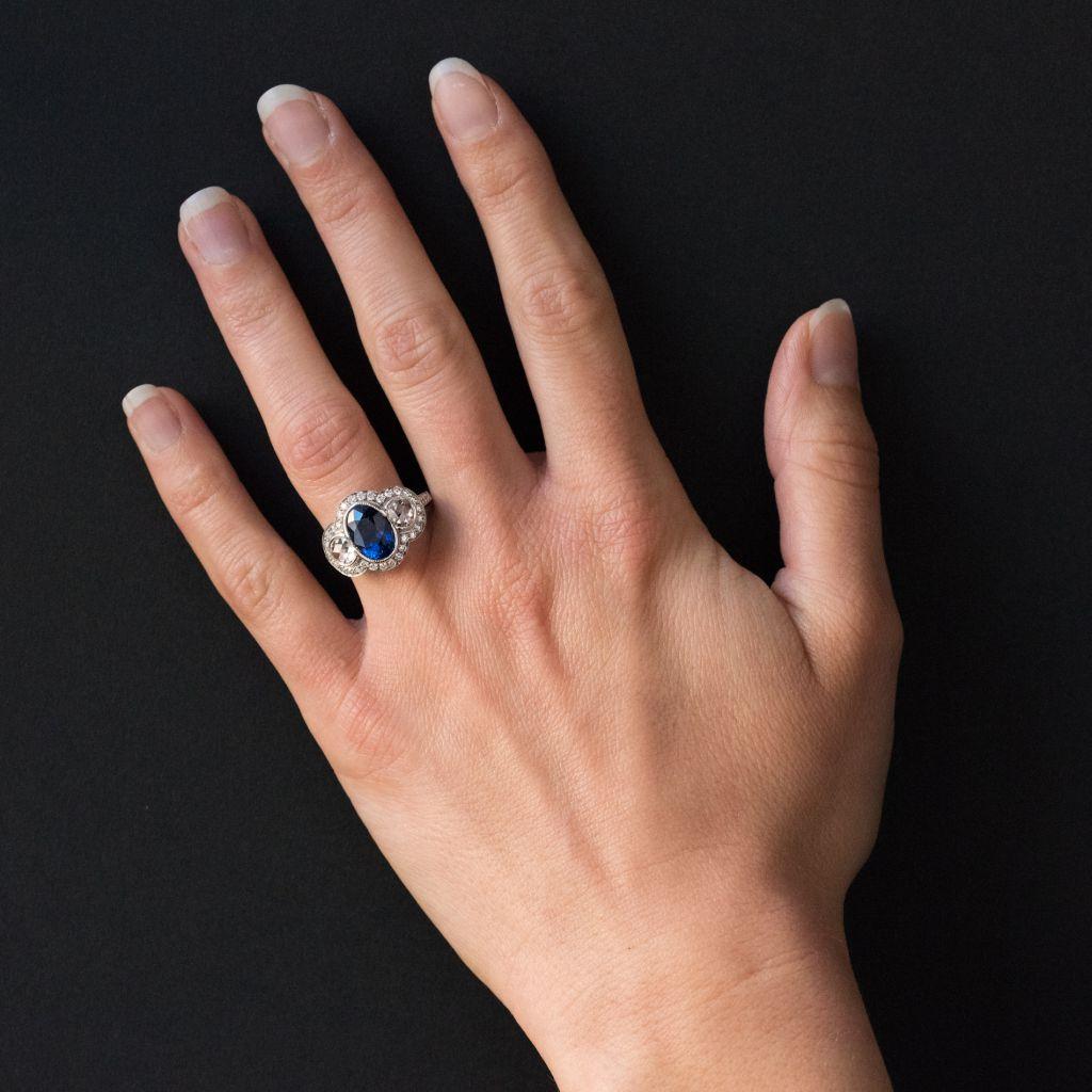 Ring in 18 karat white gold, eagle head hallmark. 
This ring features a central oval sapphire with a beaded setting shouldered by a rose cut diamond at each side, surrounded with claw set diamonds. The shoulders of the ring band are set with 2 x 3