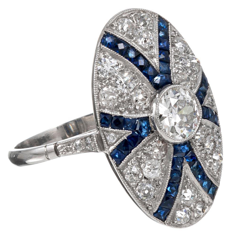 Created by hand in classic art deco tradition, this intricate platinum ring features a .61 carat old European cut diamond at its center with a detailed celebration of additional diamonds and blue sapphires arranged in an artful frame. The center