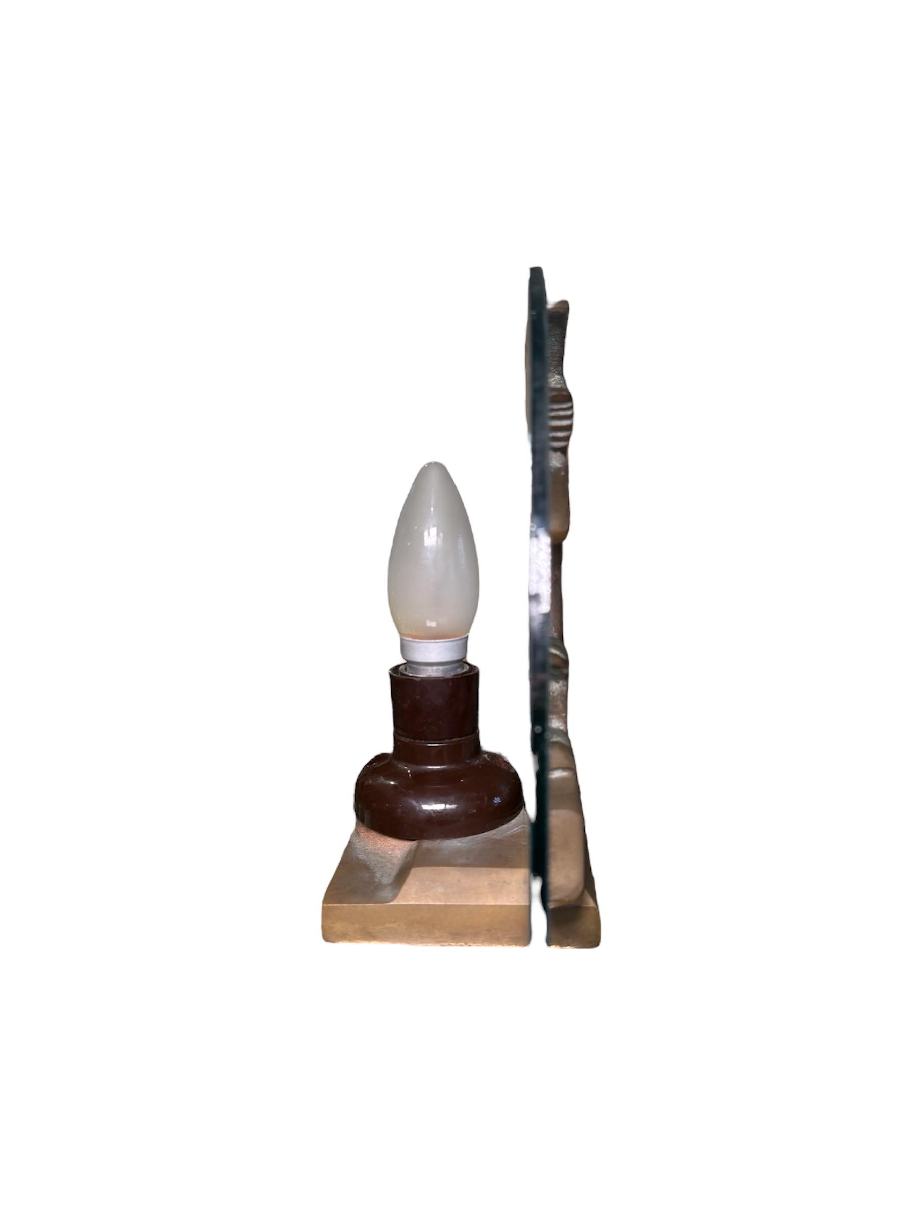 This is an Art Deco Style Table Lamp. It depicts a small table lamp with a metal sculpture of Diana the hunter and her dog supported by a metal rectangular base. Behind the sculpture, there is a half moon glass and in the back of it, it is found the