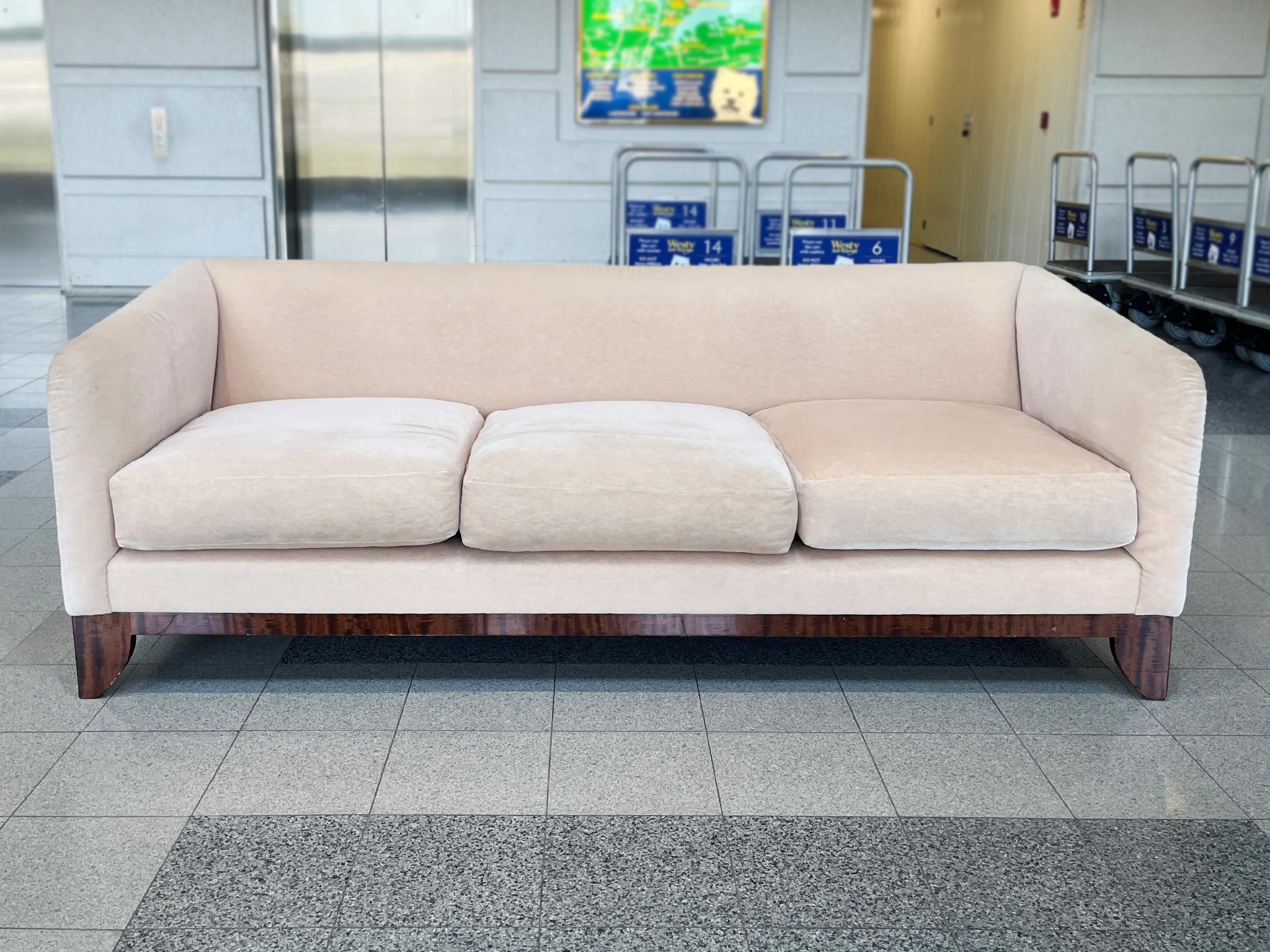 Smooth lines and full, rounded forms characterize the design of this comfortable Art Deco style sofa by J. Robert Scott. Made in the late 20th Century, the sofa is upholstered in a pale pink mohair. Its base and legs are hardwood with a dark burl