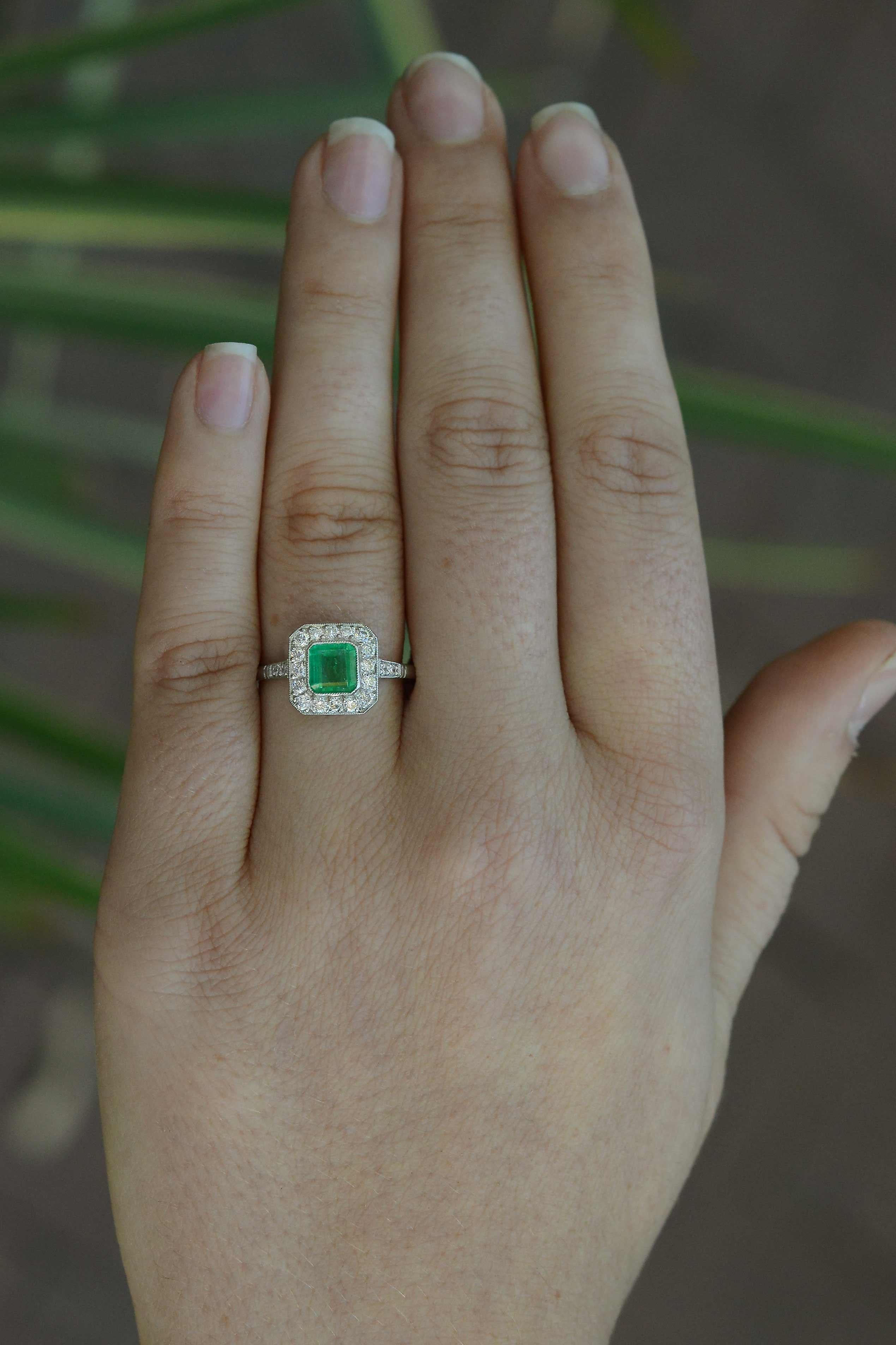 A breathtaking grass-green Colombian emerald engagement ring wows with a striking Art Deco style. The platinum setting features a gemstone that glows with a light from within its soul. Floating in a bezel setting and surrounded with a dainty halo of