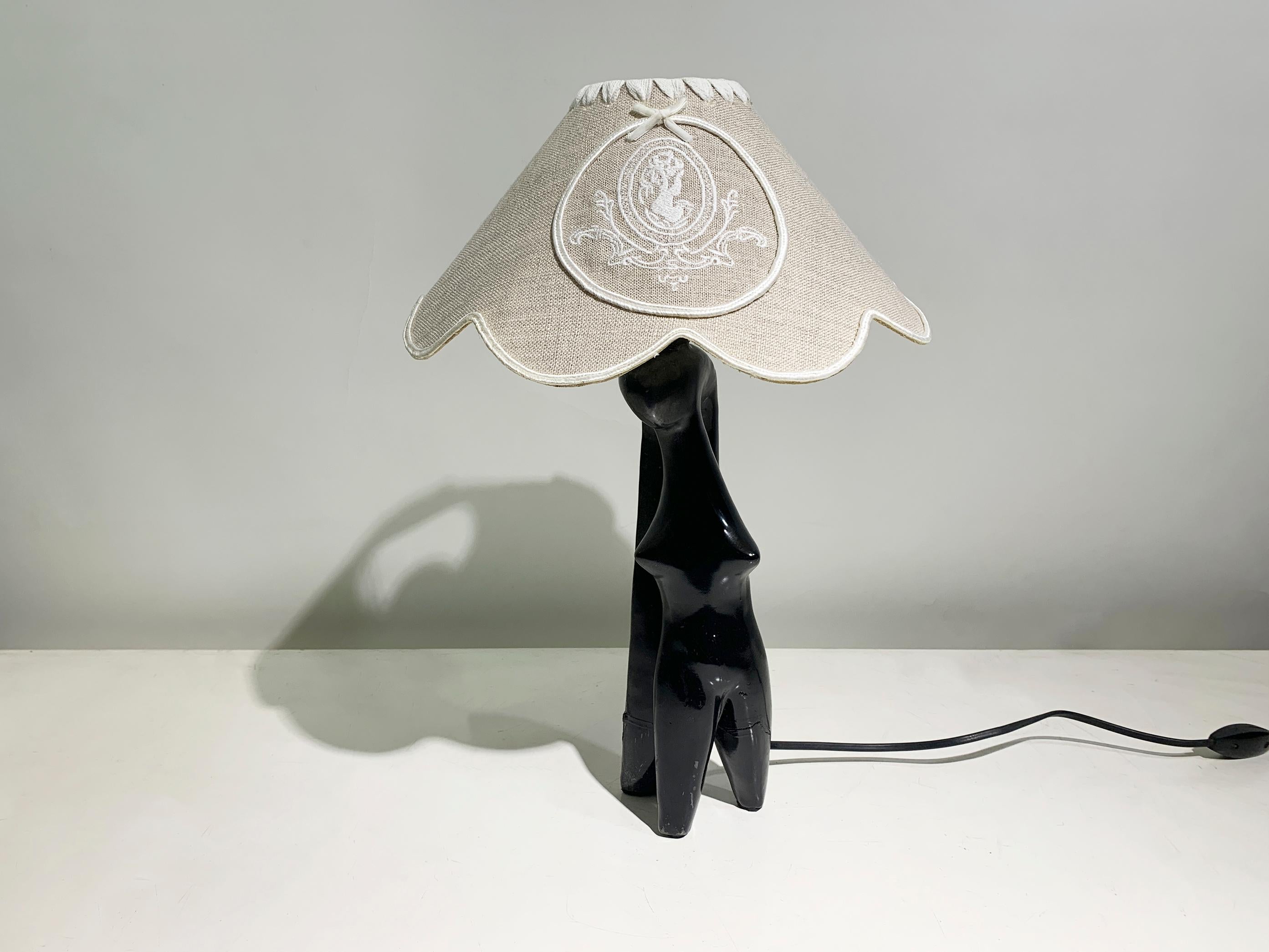 A captivating French table lamp from the 1970s, featuring a stylized black lacquered female figure in the Art Deco style. The sculptural base elegantly supports a wide, scalloped linen lampshade adorned with an embroidered scalloped shade, adding a