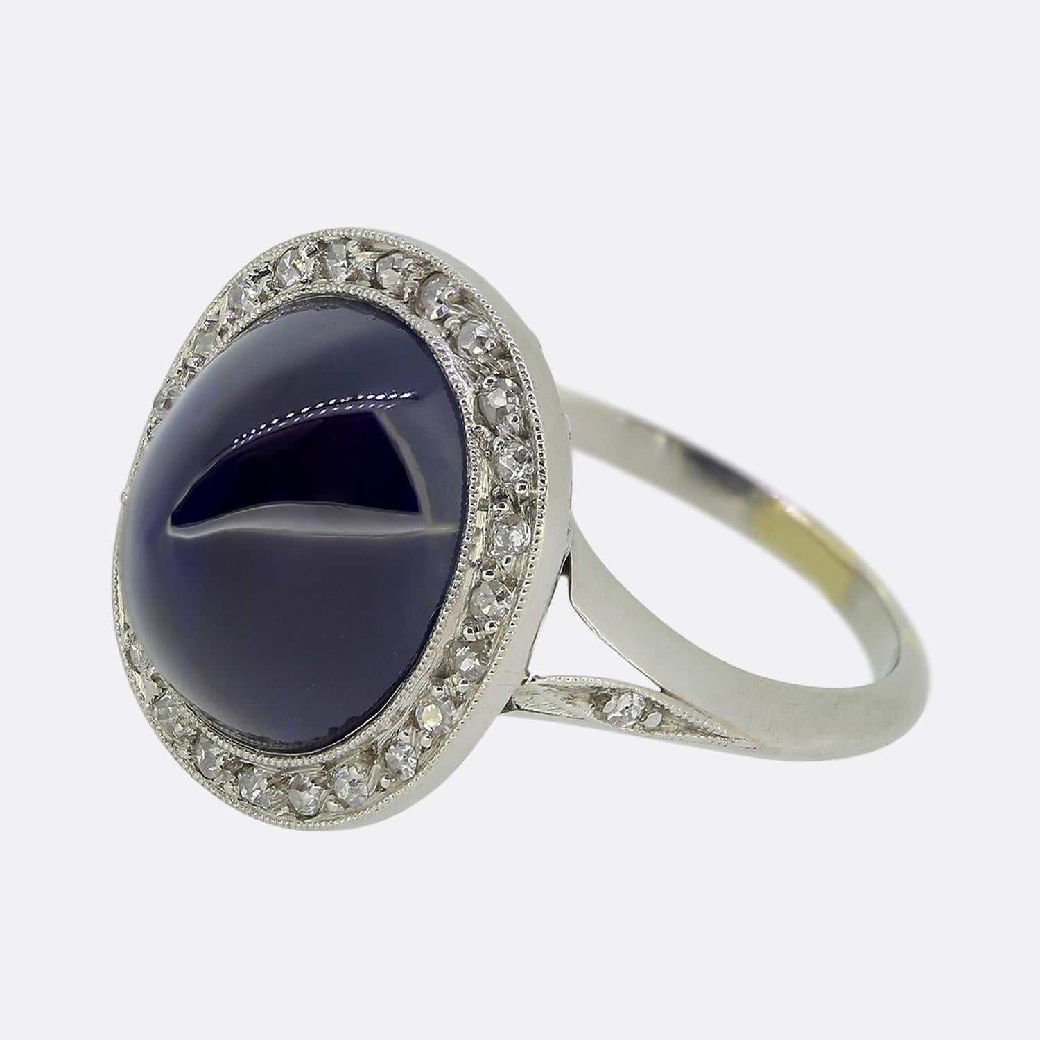 Here we have a gorgeous cluster ring crafted during the Art Deco movement. At the centre of the face we find a captivating sugarloaf cabochon sapphire possessing a deep navy blue colour tone. This principal stone sits proud whilst being surrounded