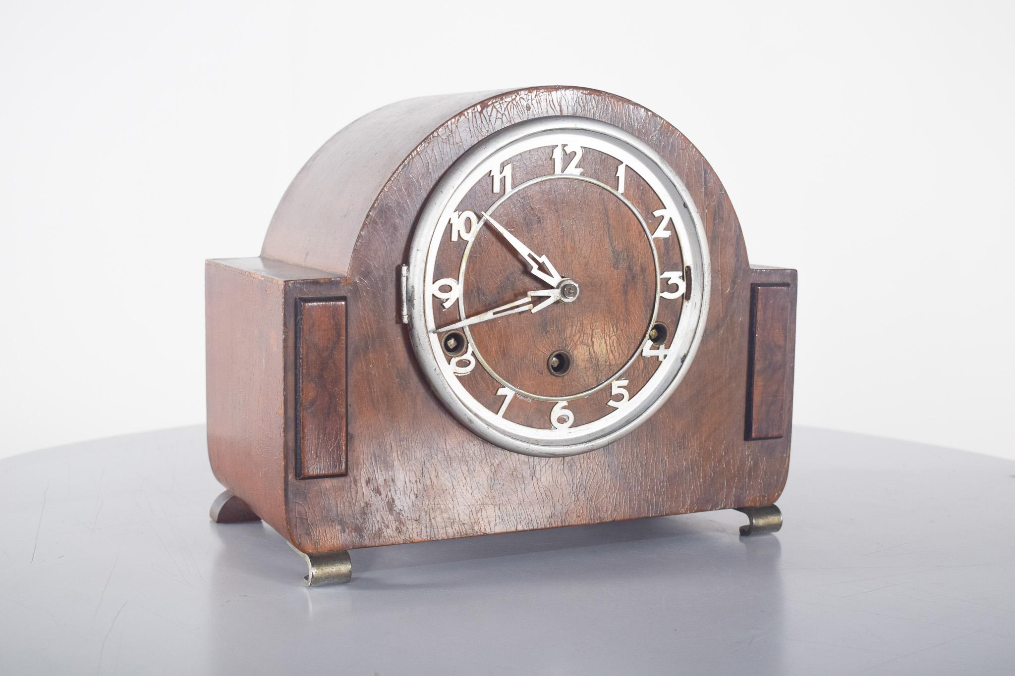 Art Deco Tischuhr. Aus dunklem Holz, das an traditionelle Kaminsimsuhren erinnert. Die halbrunde Struktur verbindet sich mit den geometrischen Details an den Seiten und schafft ein Gleichgewicht zwischen Schlichtheit und Raffinesse.
Das Zifferblatt