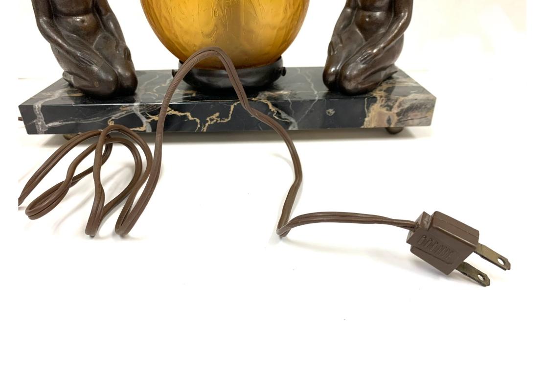 Art deco table lamp, possibly by Nuart, comprised of a textured amber globe light flanked by two female nudes and surmounted on a variegated marble base with ball feet. The globe is fastened by three tightening screws (one missing) and operates with