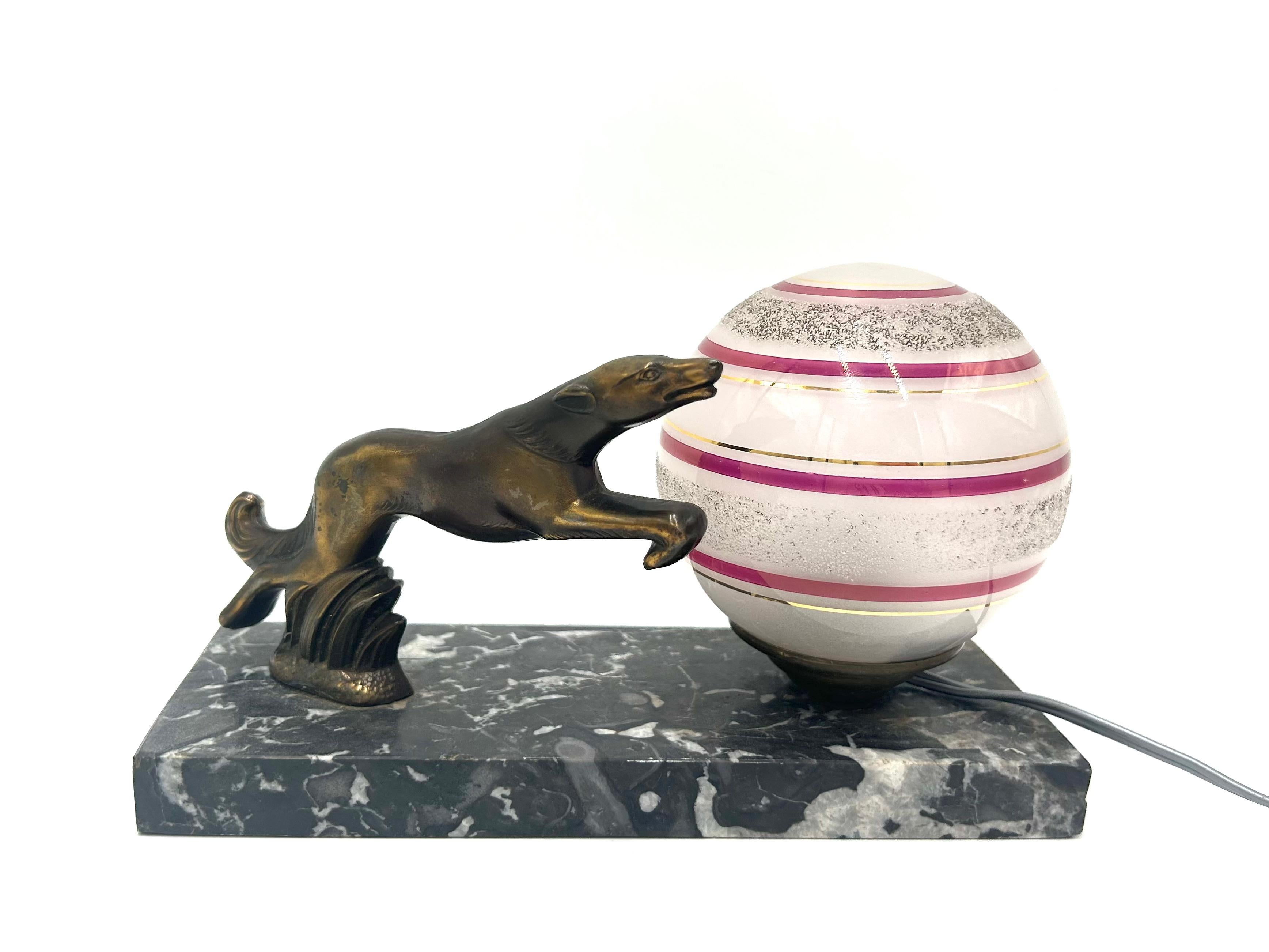 Lampe de table Art Déco. Il se compose d'un socle en marbre, d'un abat-jour en verre en forme de boule et d'une figure de chien courant (lévrier). Ampoule de type B22 - plus petite taille. Une pièce est incluse dans le set. La lampe a été fabriquée