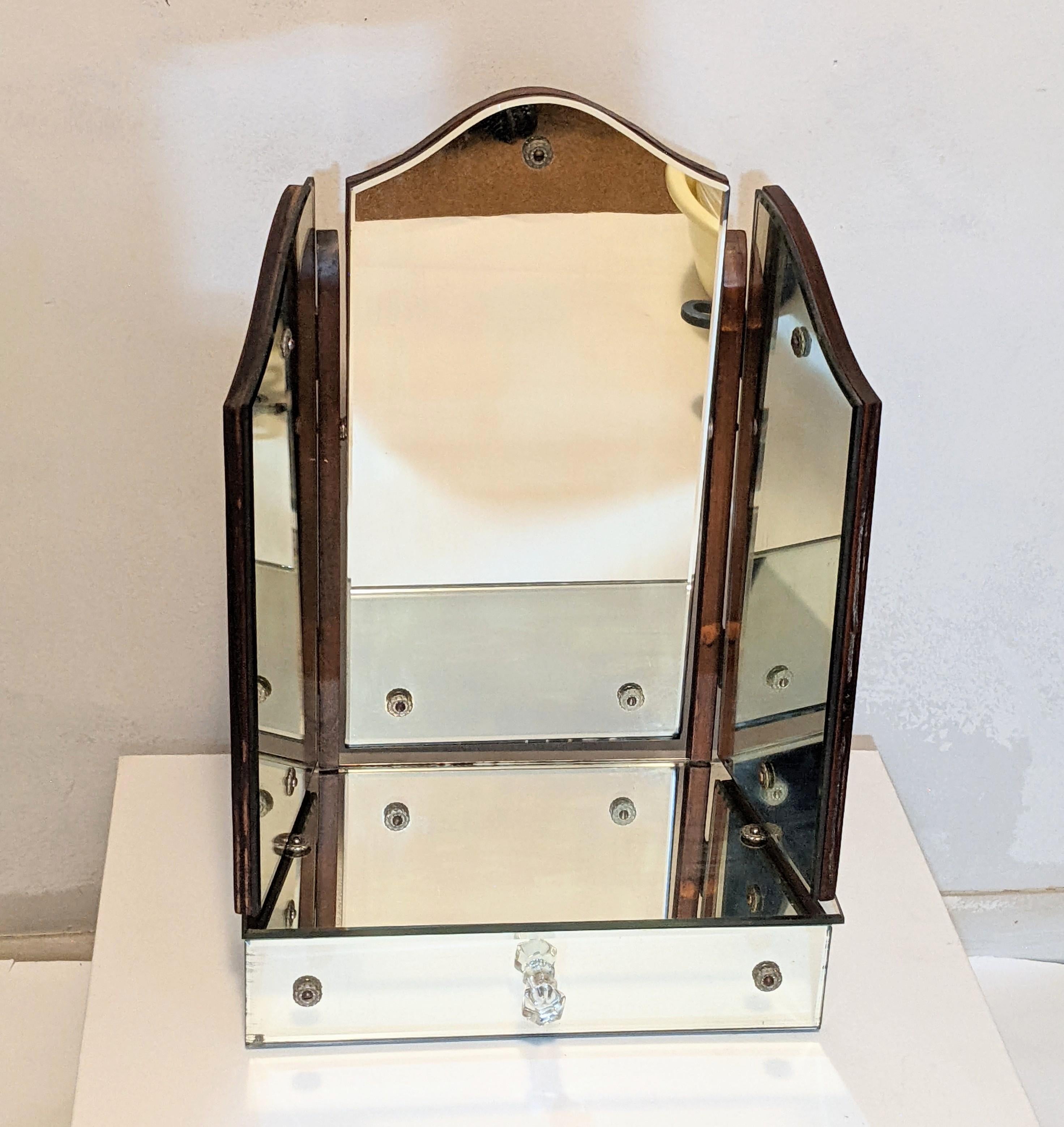 Art Deco table top dressing mirror with drawer from the 1930's. Backed in wood with 3 mirrors, the center one tilts up and down while the side panels tilt outwards. 
Decorative fluted crystal pegs anchor each of the mirror panels and a molded glass