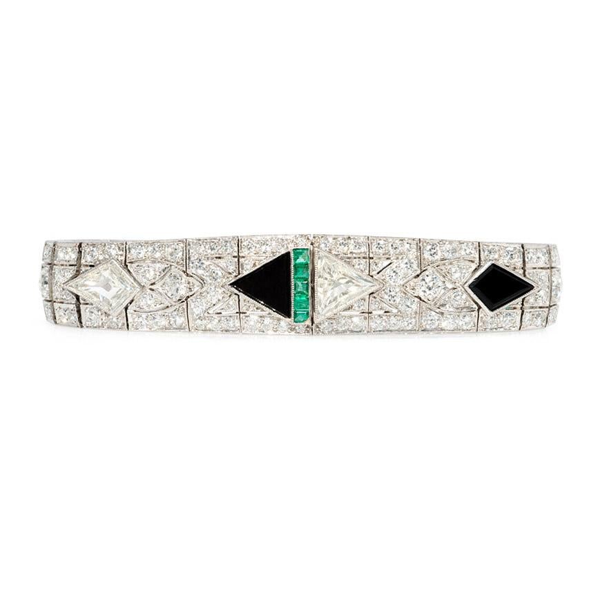 An Art Deco diamond strap bracelet of tapered design centering on a triangular-cut onyx and a diamond trilliant with alternating bullet-shaped cuts and calibré emeralds throughout, in platinum.  Numbered #549.  Atw 9.06 ct. diamonds.

Approximately