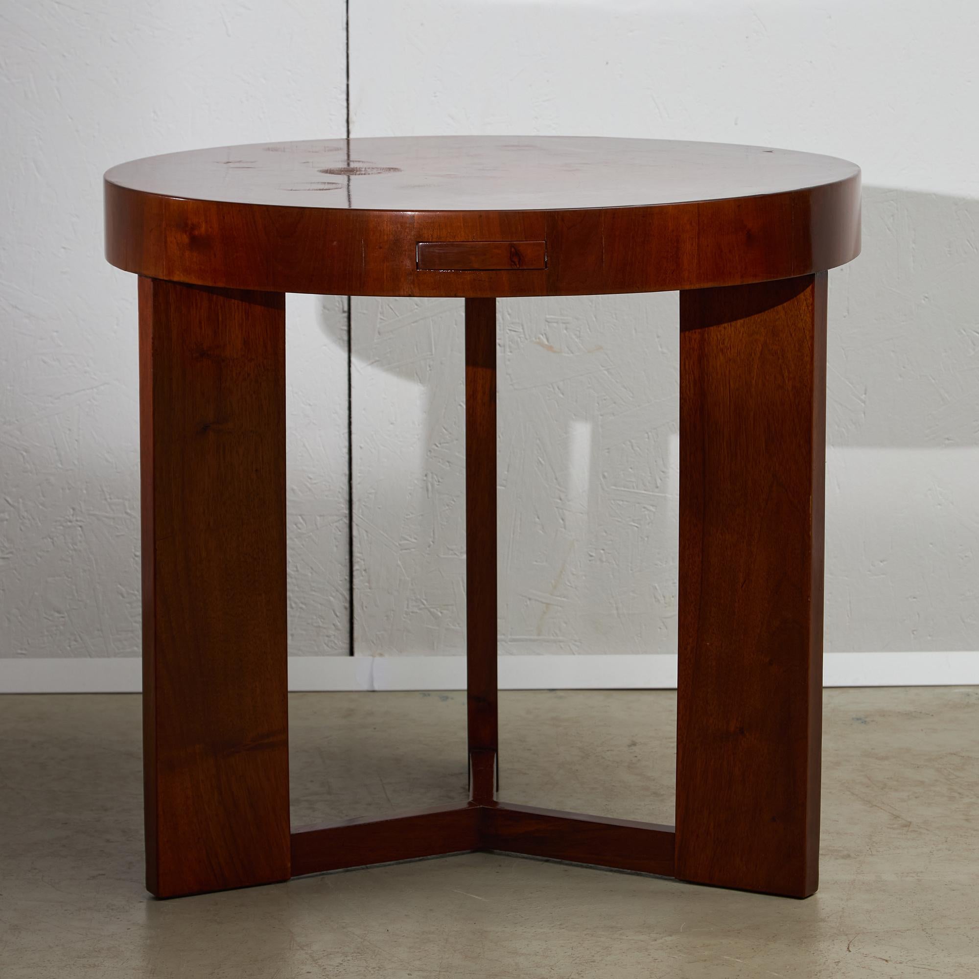 This Art Deco round table, crafted from richly grained teak, embodies the refined yet practical design sensibilities of the early 20th century. The tabletop is supported by three interlocking legs, a structural detail that provides stability while