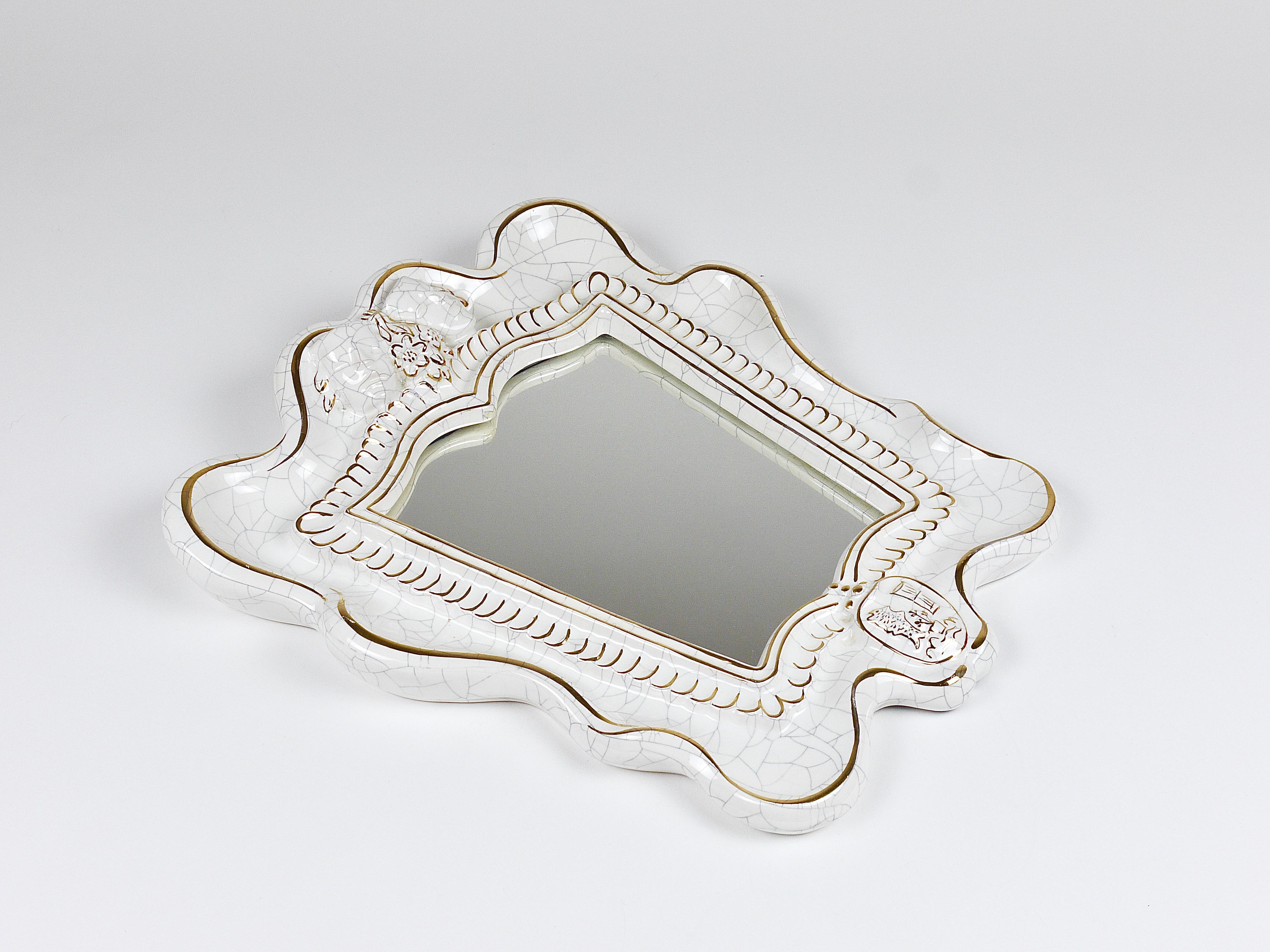Charmant miroir mural de style Art Deco du milieu du siècle, fabriqué dans les années 1950 par Gmundner Keramik en Autriche. Le miroir présente un cadre en céramique/terracotta en forme de bouclier avec une glaçure craquelée blanche. Deux masques de