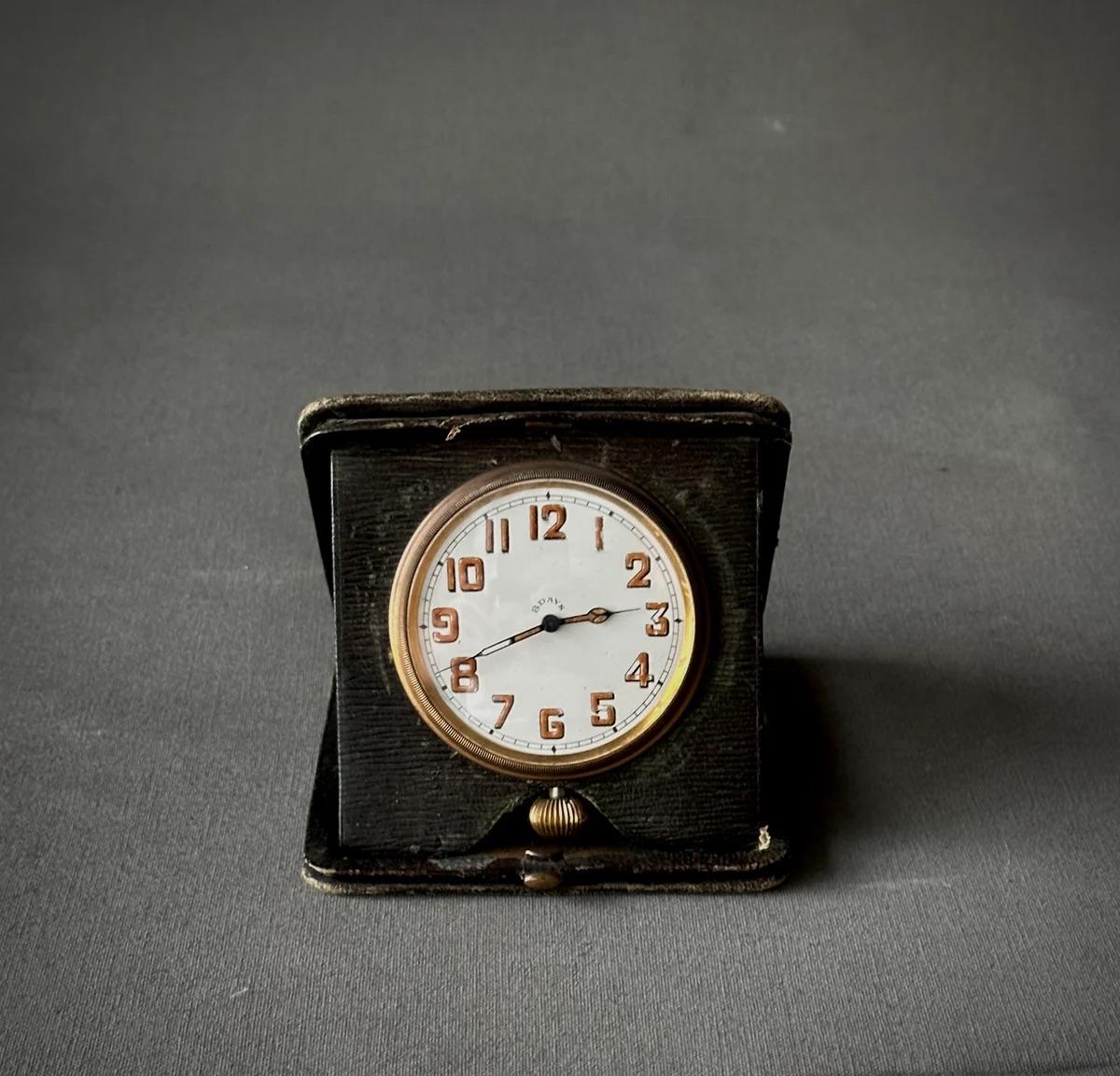 This Art Deco travel clock from England, encased in a dark green textured leather foldable case, radiates elegance and practicality. Its white ceramic face features delicately hand-painted gold and black numerals, offering a glimpse into the