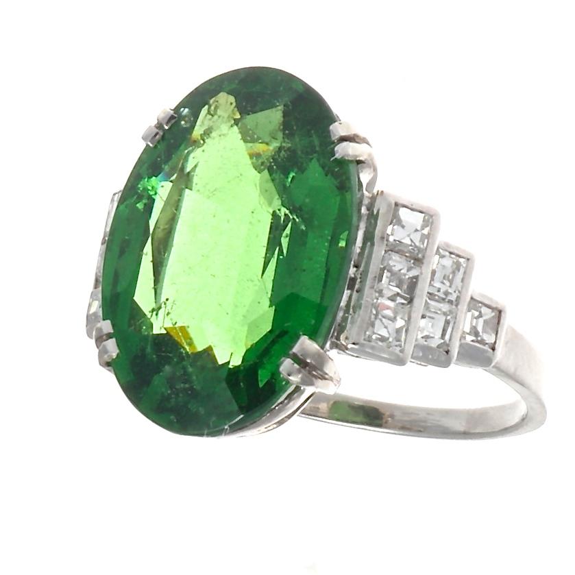 A sparkling vivd green tsavorite garnet is well matched with diamonds, set in a classic Art Deco setting. The 6.90 carat tsavorite garnet is prong set in platinum and is accompanied by 12 Carre cut diamonds that weigh approximately 0.60 carats and