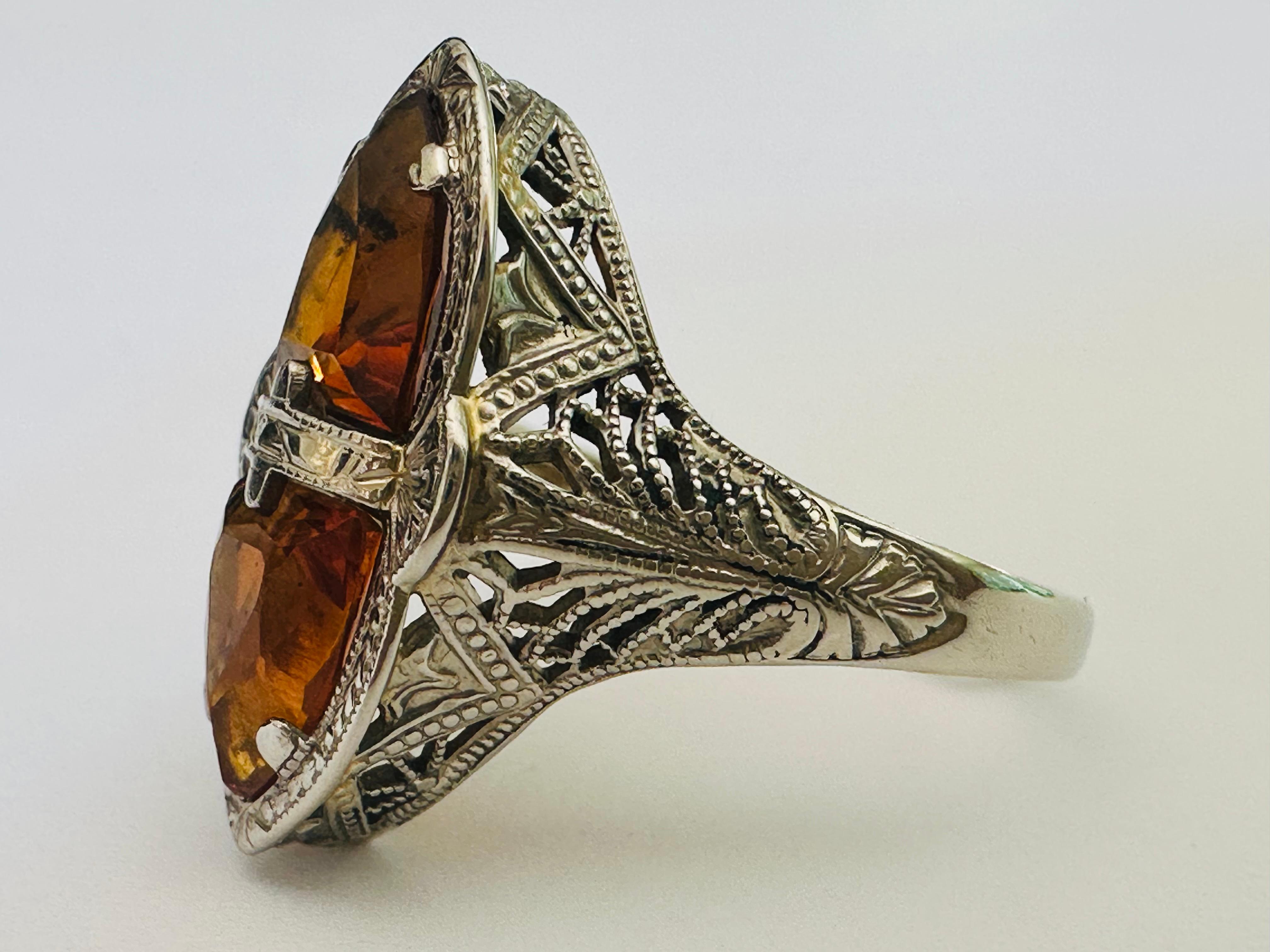 Deux citrines naturelles de couleur orange dorée, taillées en trillion, rayonnent sur cette bague Art déco, vers les années 1930. Les pierres vibrantes se trouvent au sommet d'une galerie de filigranes en dentelle conçus avec art. Serti en or blanc