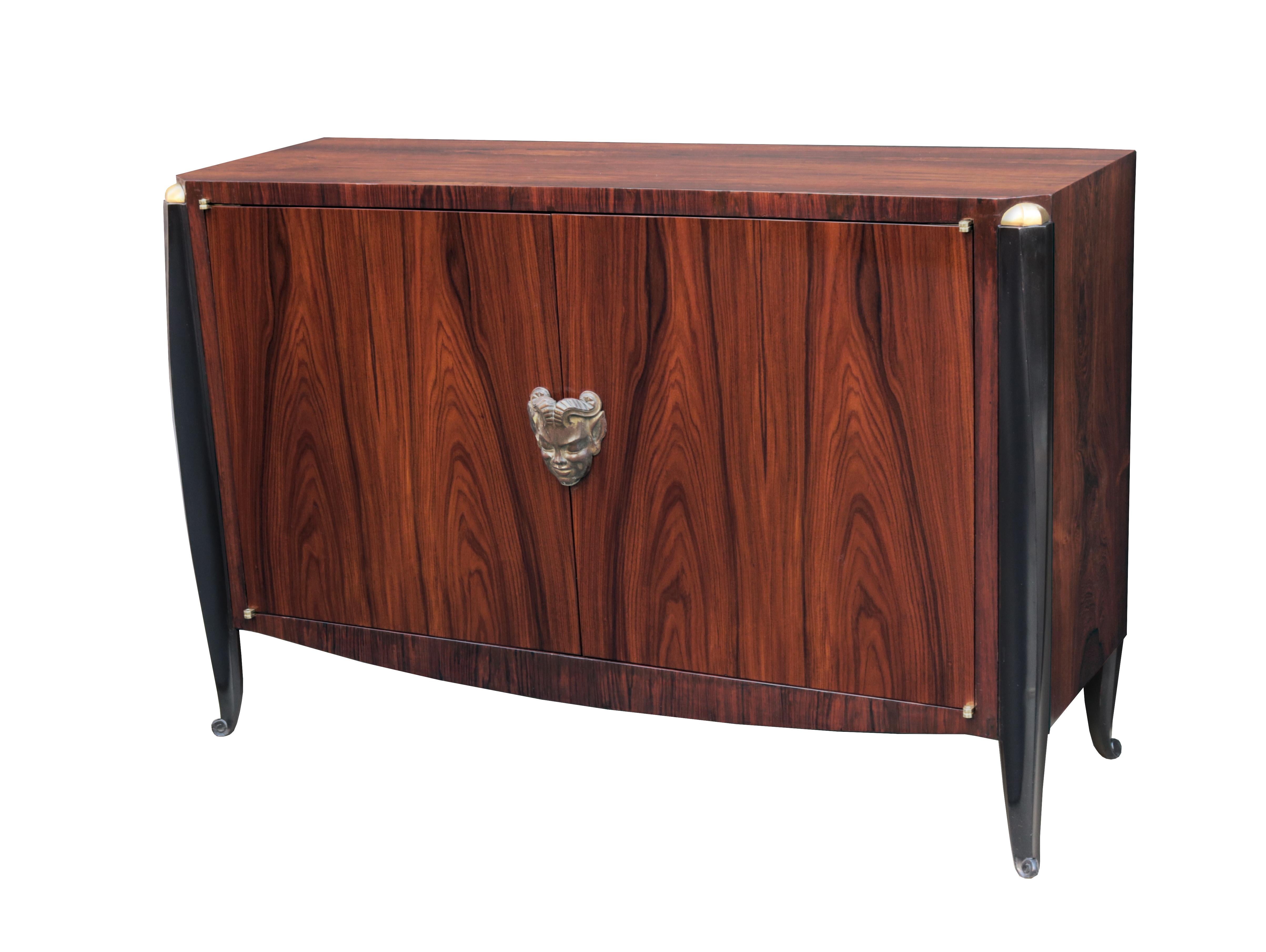 An Art Deco two door cabinet attributed to Jean Pascaud.
Rosewood with ebonized legs, giltwood details, central carved giltwood face detail.
